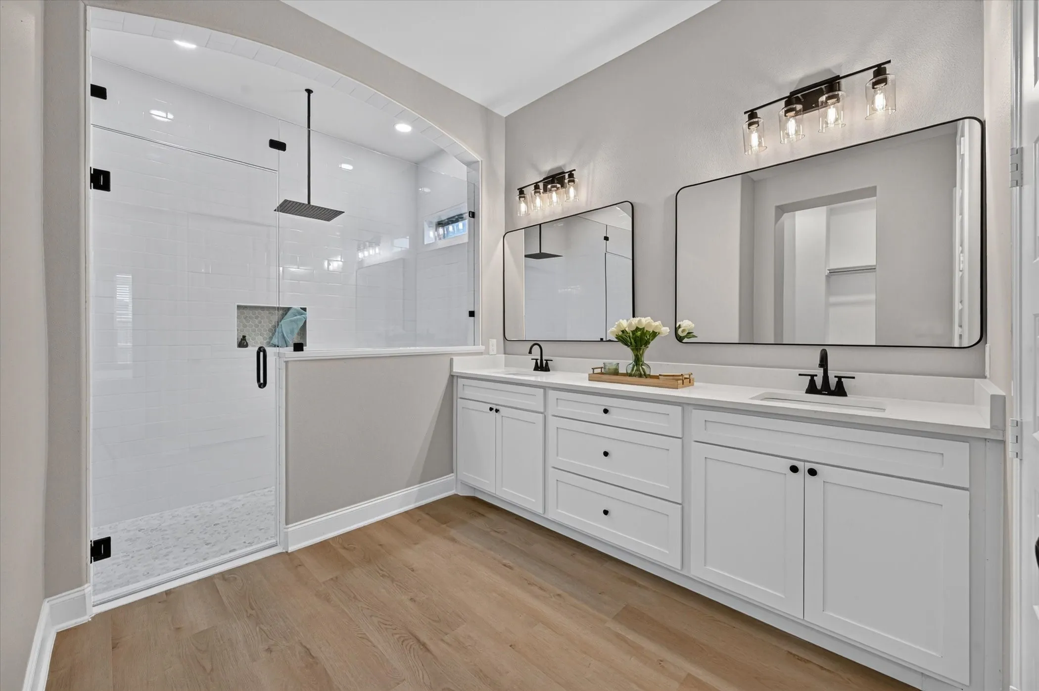 Spacious bathroom featuring a frameless glass shower enclosure, dual vanity with white cabinetry, black framed mirrors, and wood-finish flooring