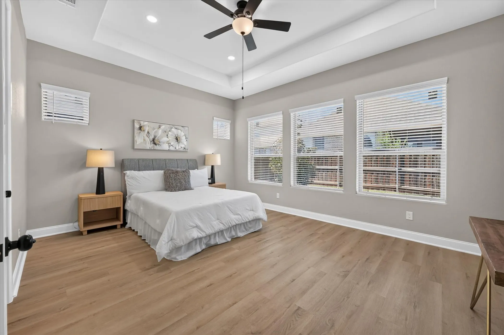 Tray ceiling with recessed lighting and ceiling fan, three windows with blinds, wood-finish flooring, and light-toned walls