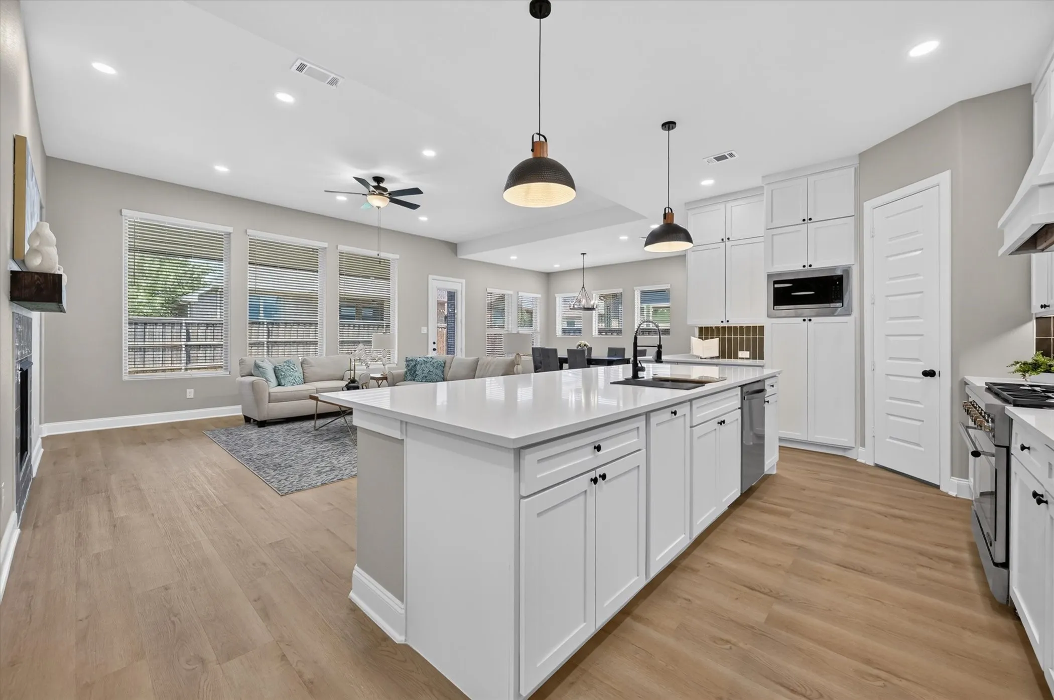 Spacious open-concept living area featuring wood-finish flooring, a large kitchen island with a sink, white cabinetry with dark hardware, a built-in microwave, and recessed lighting