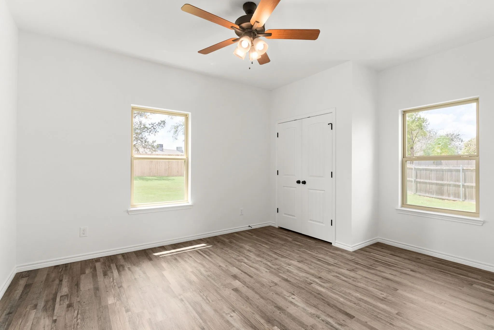 Bright interior with wood-finish flooring, two large windows, a ceiling fan with integrated lighting, and a double-door closet