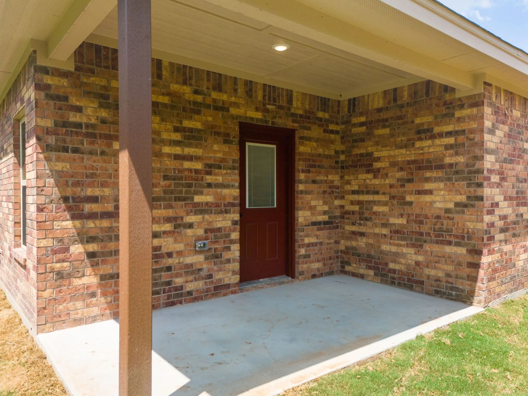 Covered patio with a concrete surface, multicolored brick exterior, recessed lighting, and a red entry door with a frosted glass insert