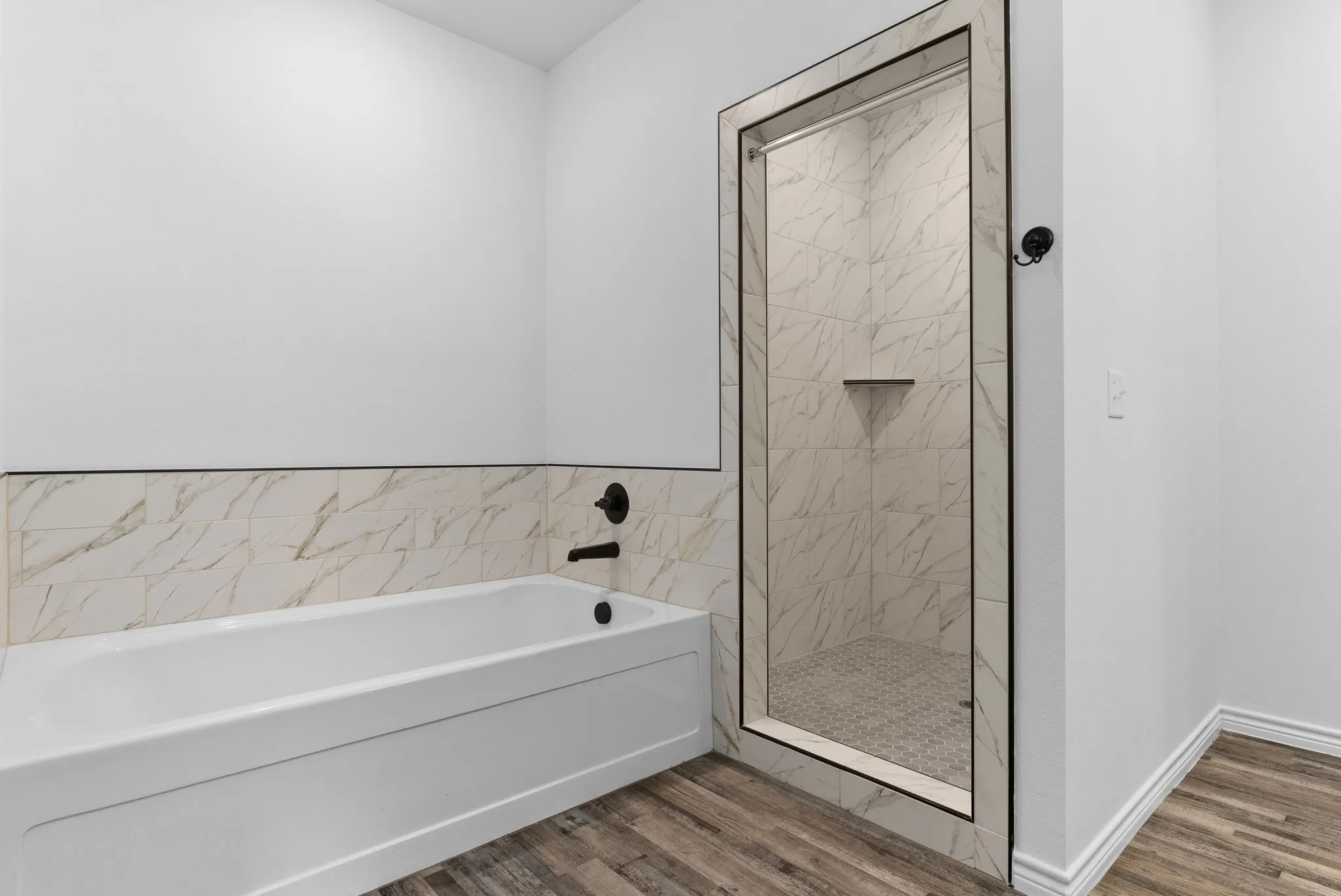 White soaking tub with matte black fixtures, an enclosed shower featuring marble-look tile, and wood-finish flooring