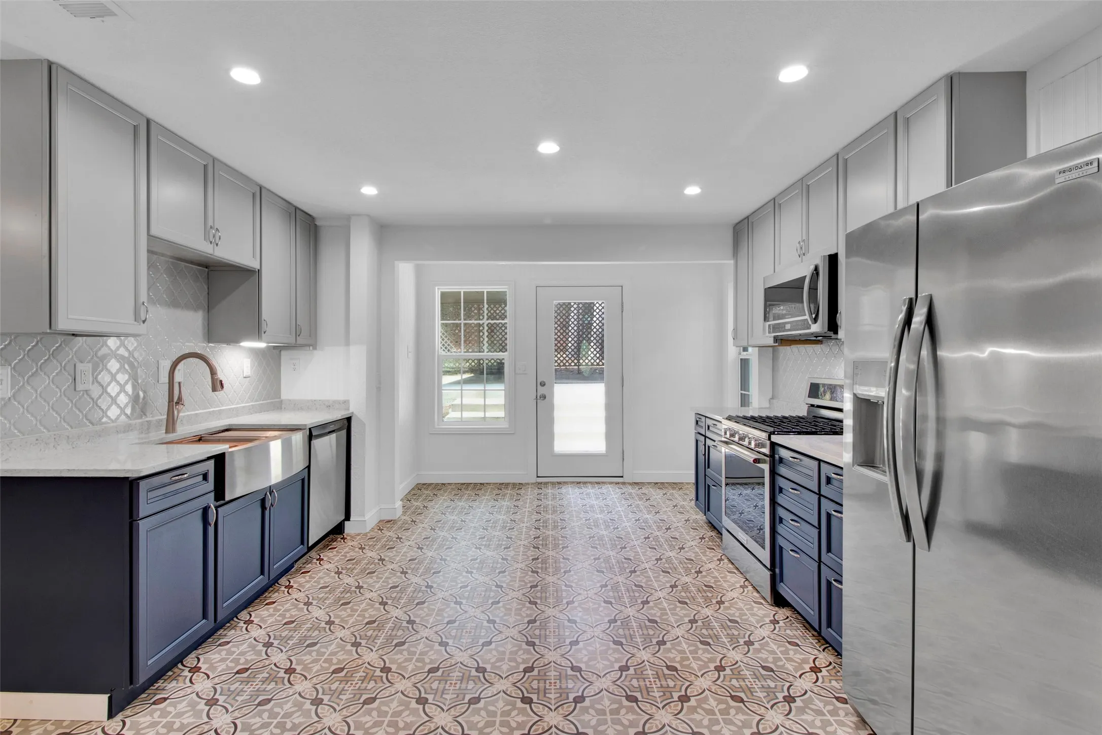 Contemporary kitchen featuring two-tone cabinetry, stainless steel appliances, a farmhouse sink with a bronze-finish faucet, and patterned floor tiles