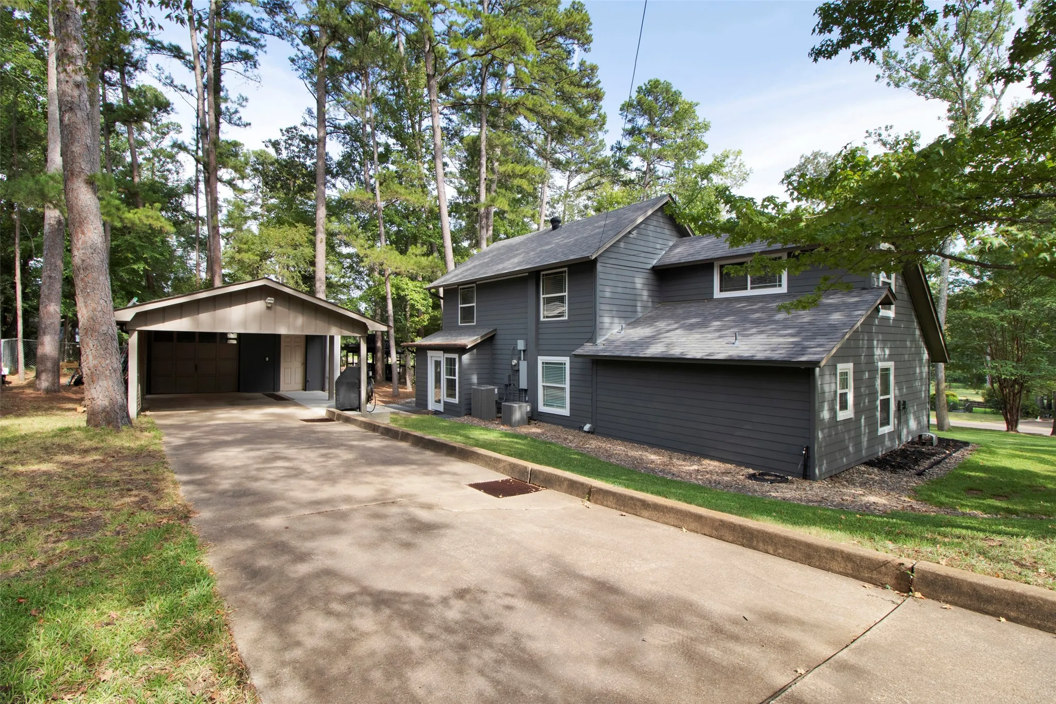 Gray siding exterior with white window trim, mature trees, and a detached carport