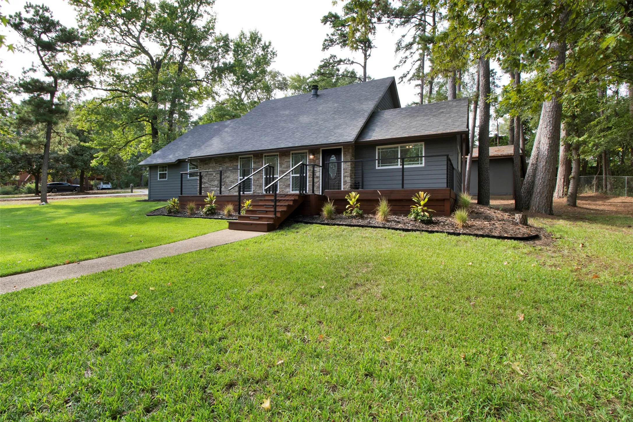 Exterior featuring gray siding, stone accents, a dark shingle roof, a wood-finish deck with dark railings, and a concrete pathway