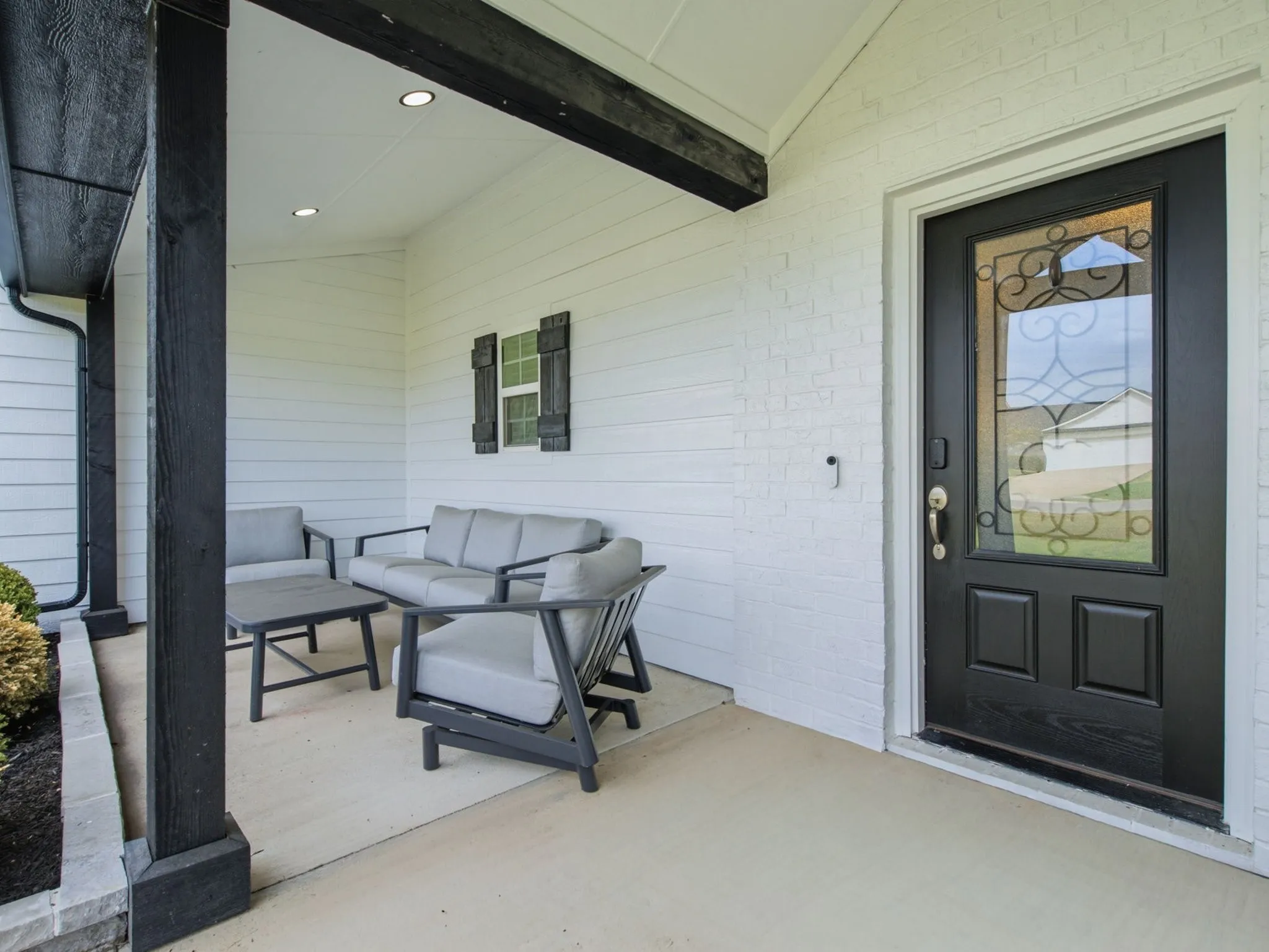 Covered front porch featuring a dark-stained wood beam and columns, white brick and siding exterior, recessed lighting, and a dark-paneled entry door with decorative glass