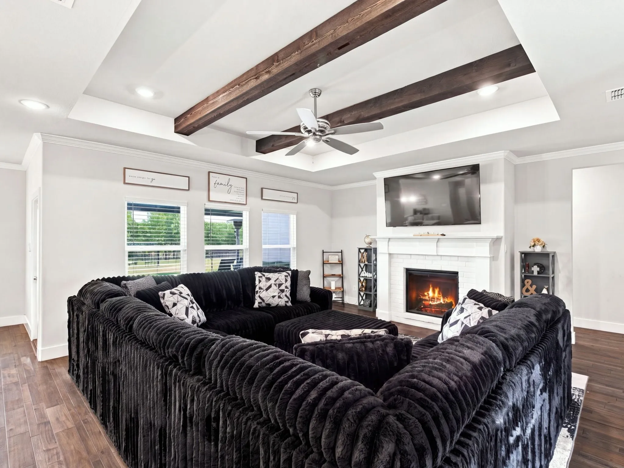 Tray ceiling with exposed wood beams, ceiling fan, wood-finish flooring, white brick fireplace with mantel, and recessed lighting