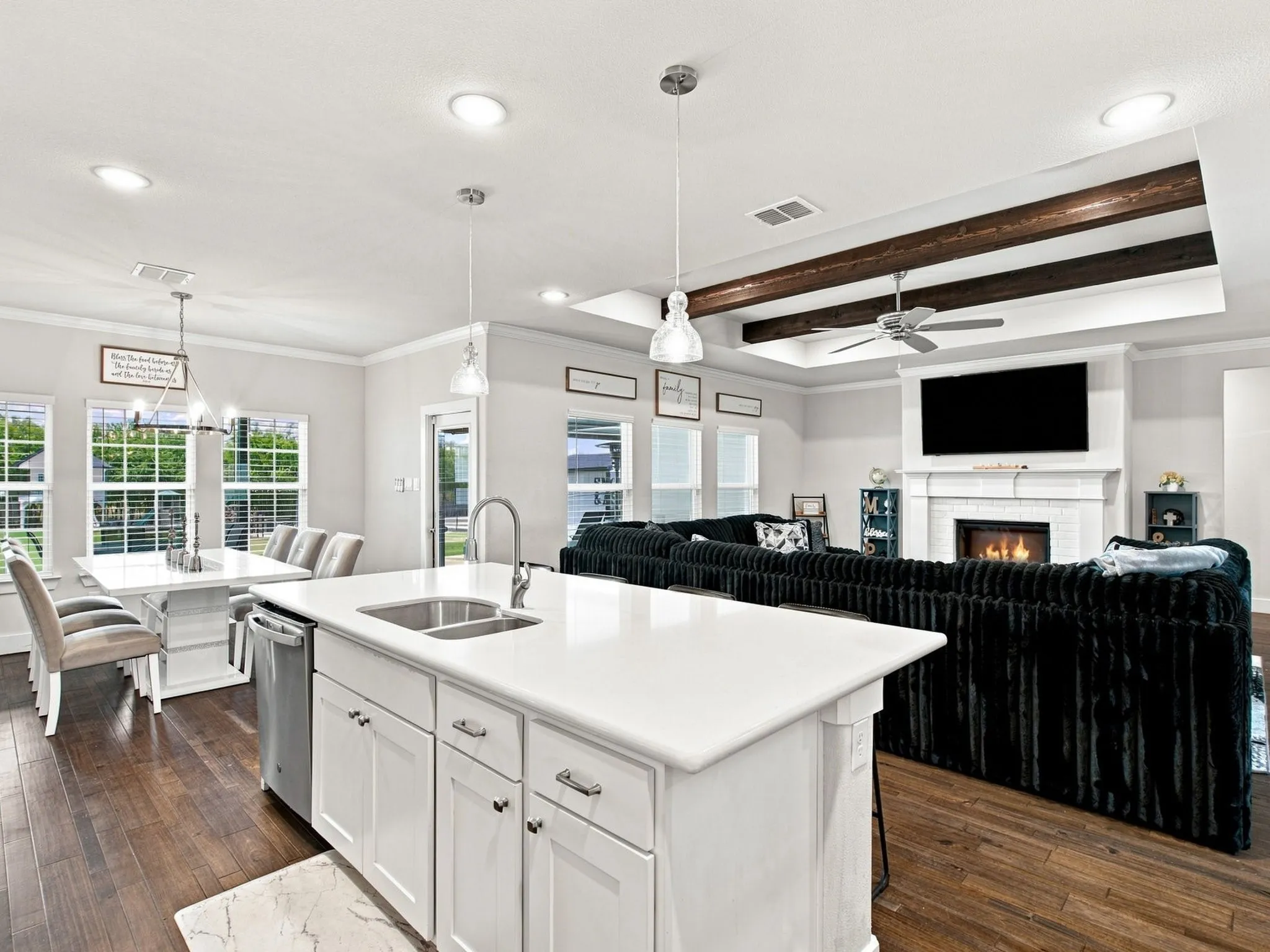 Open-concept living space with wood-finish flooring, a kitchen island with integrated sink, recessed lighting, and a living area featuring a fireplace with a mantel and exposed ceiling beams