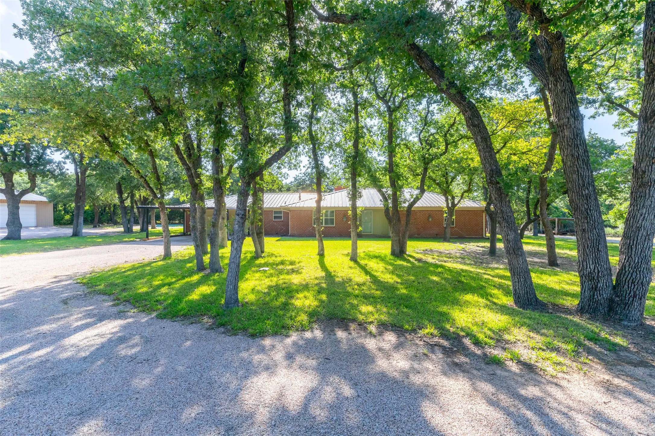 Gravel driveway leading to a brick residence with a metal roof, surrounded by numerous mature trees and a separate detached structure