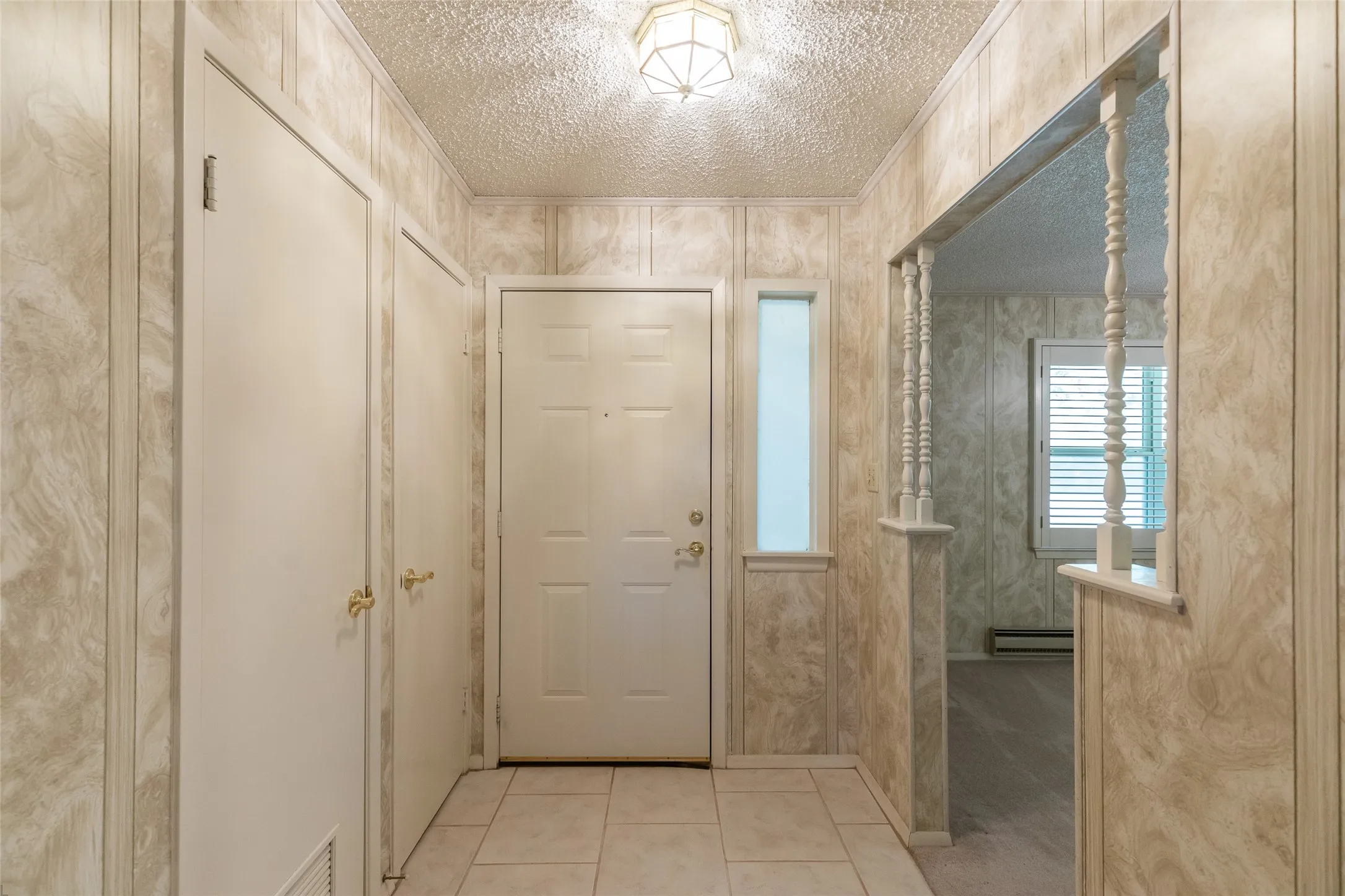 Tiled entryway featuring a panel-design door with a sidelight, two storage closets with brass hardware, and an archway opening to an interior space with shuttered windows