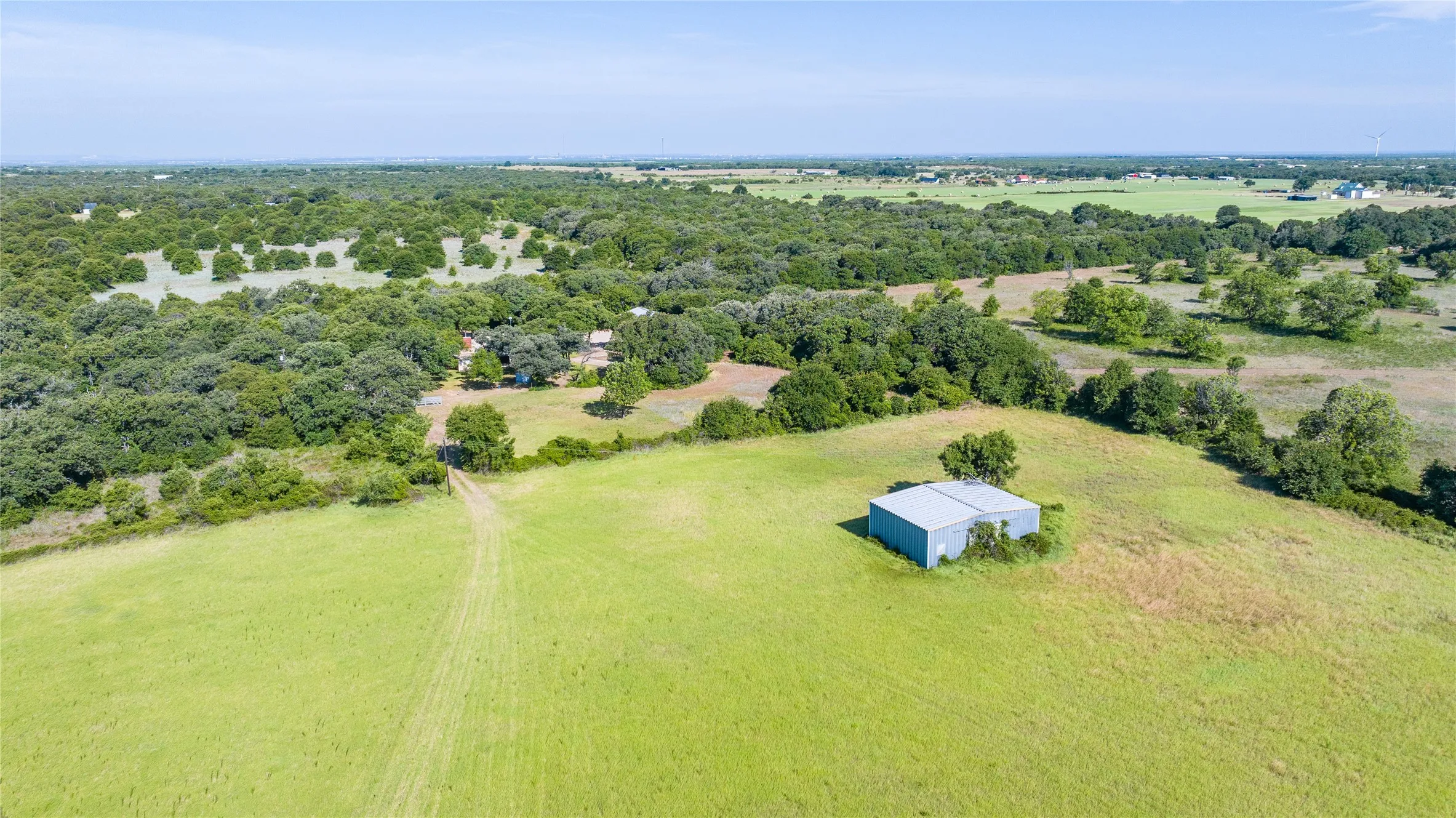Expansive green acreage featuring a detached outbuilding with a metal roof, scattered mature trees, and a distant residential structure