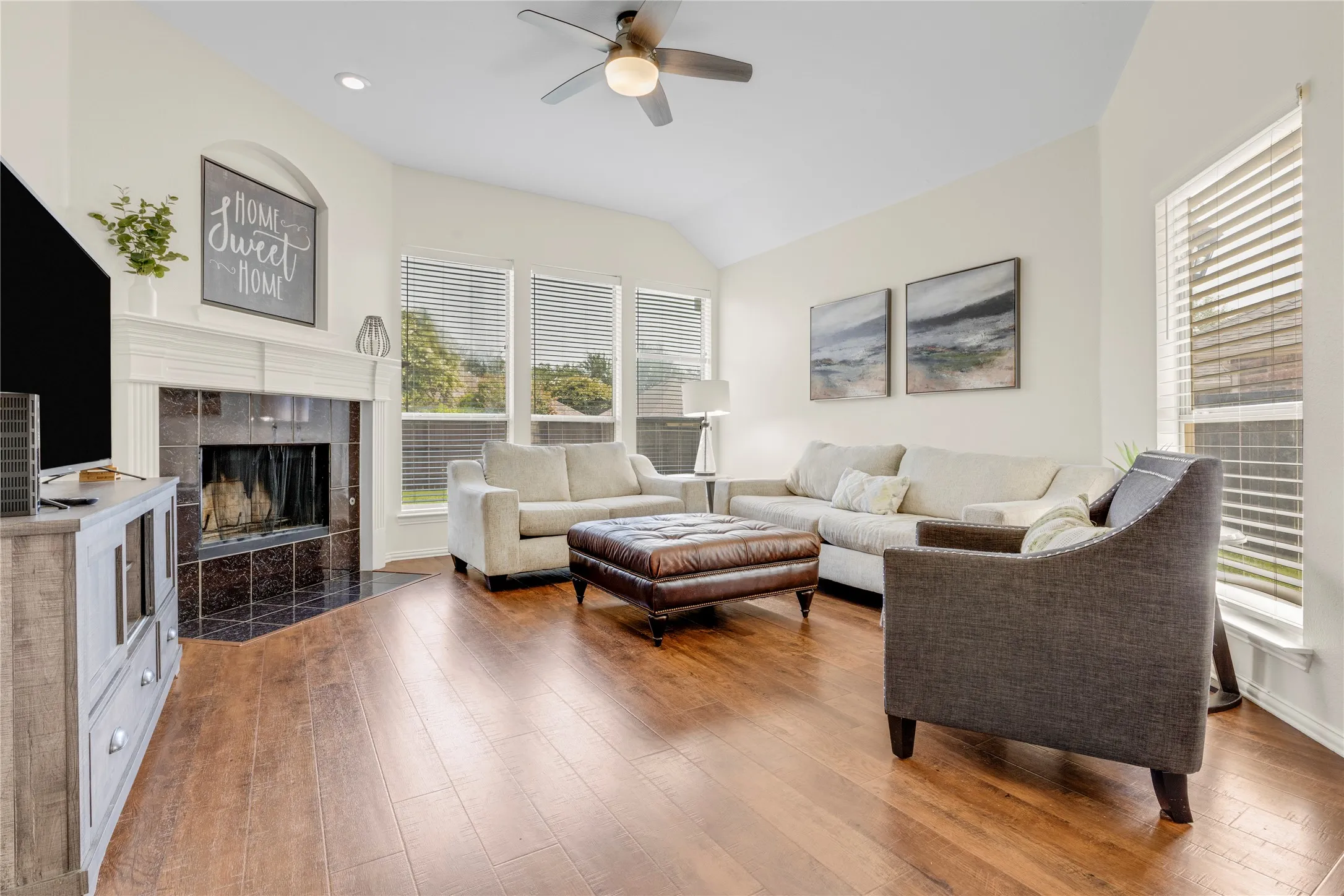Vaulted ceiling with a ceiling fan, wood-finish flooring, fireplace featuring a dark tile surround and white mantel, and multiple windows with blinds