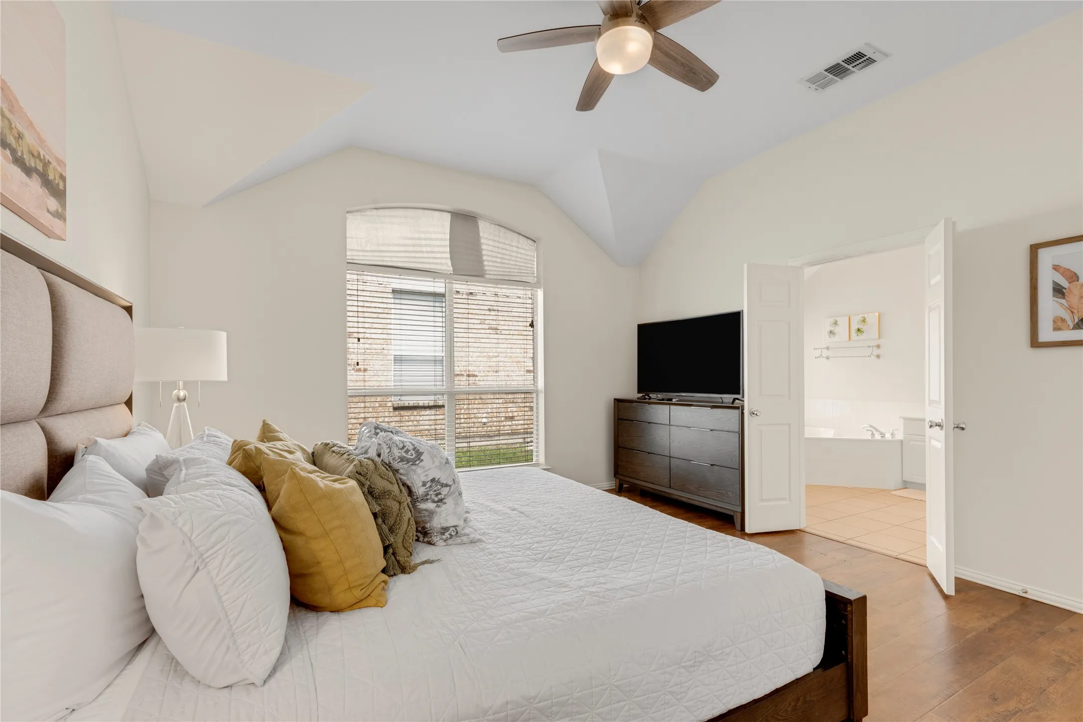 Vaulted ceiling, wood-finish flooring, arched window with blinds, ceiling fan with integrated lighting, and double doors opening to a bathroom with tile flooring and a built-in bathtub