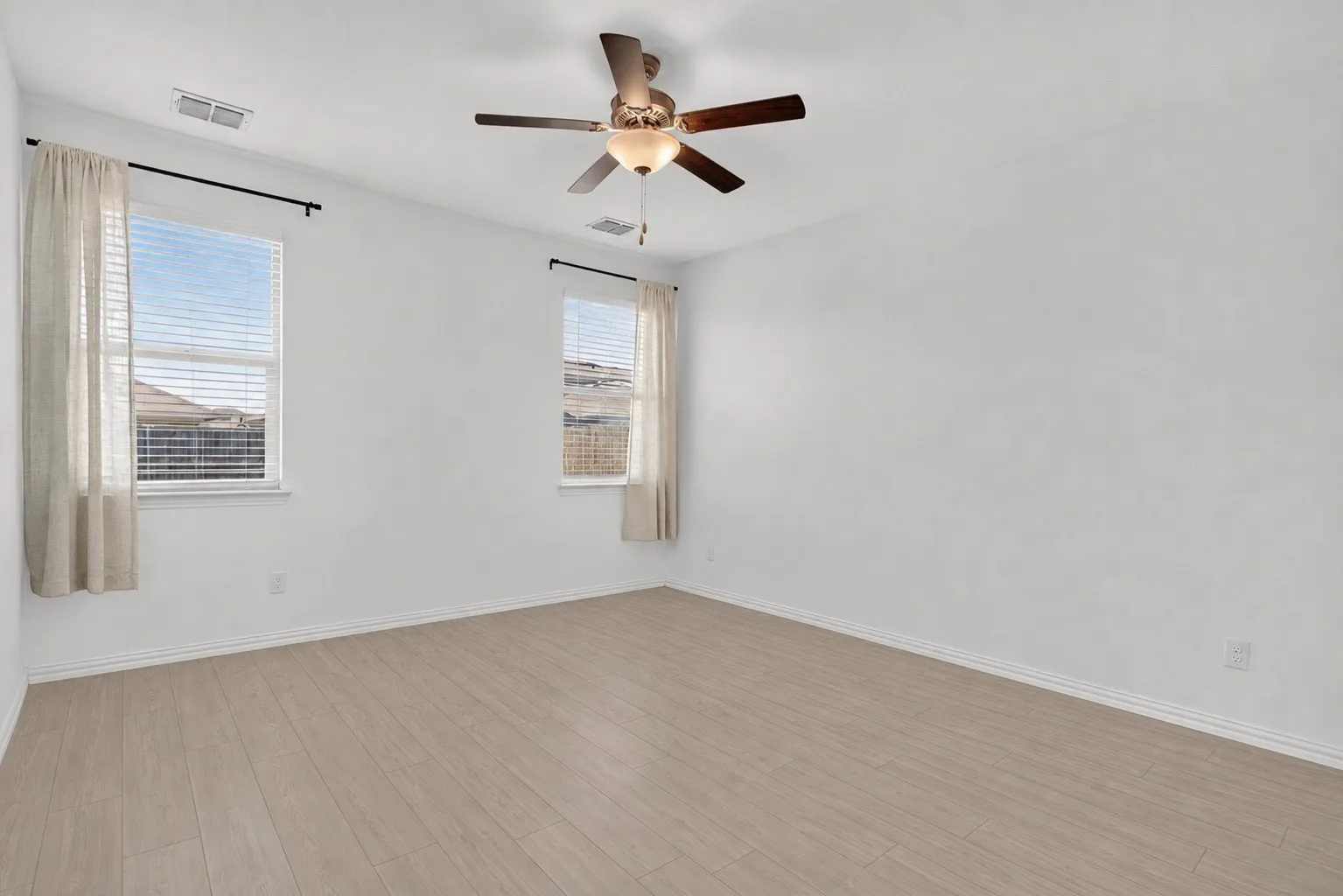 Spacious room featuring wood-finish flooring, two windows with blinds, a ceiling fan with integrated lighting, and white baseboards