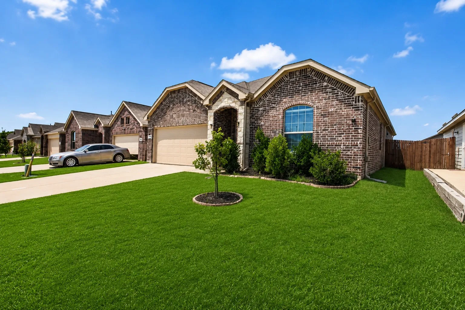 Brick exterior with arched window, integrated two-car garage, concrete driveway, and a landscaped front yard with a young tree