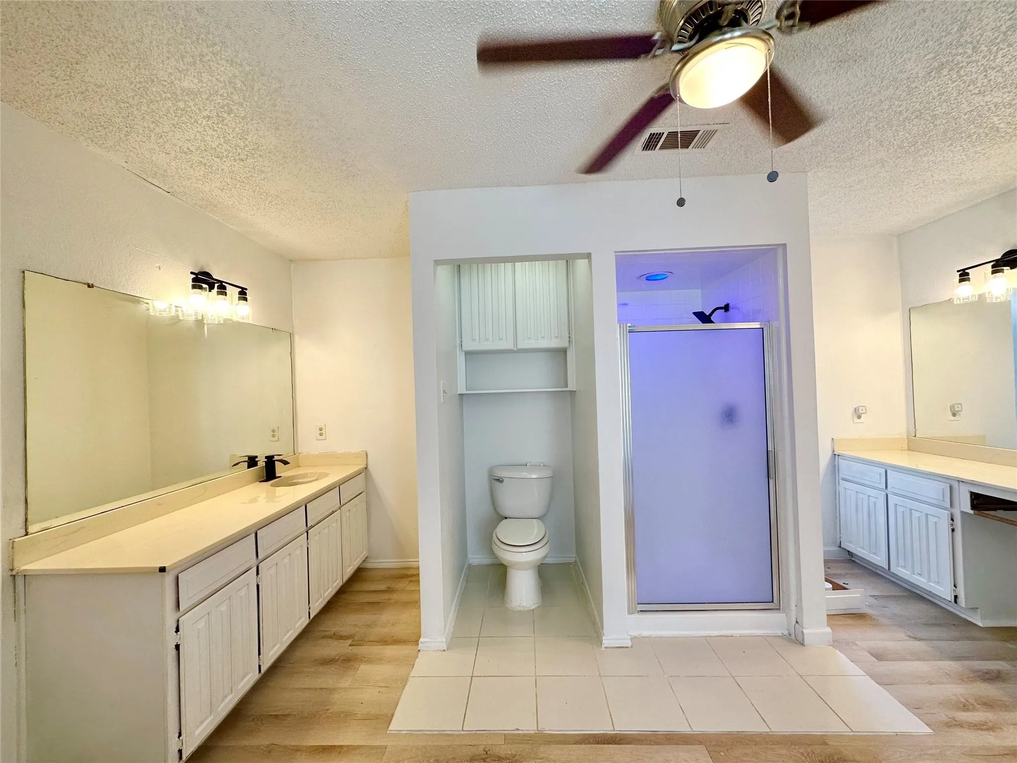Dual vanities with integrated sinks, wood-finish flooring, ceiling fan with light fixture, walk-in shower with frosted glass enclosure, and built-in toilet storage