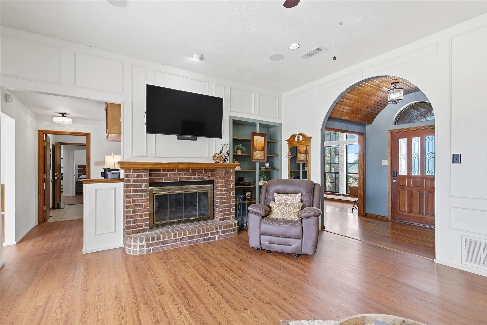 Brick fireplace with a wood mantel, wood-finish flooring, white paneling, and an arched entry with a wood ceiling