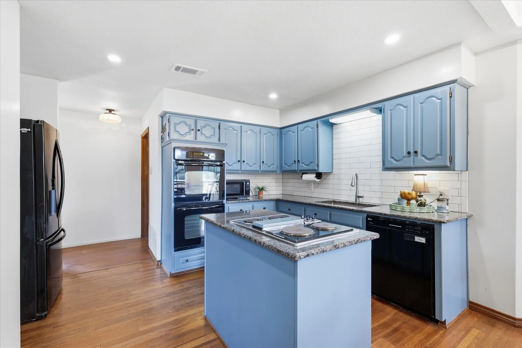 Kitchen featuring wood-finish flooring, blue cabinetry, granite-style countertops, white subway tile backsplash, and a black appliance package