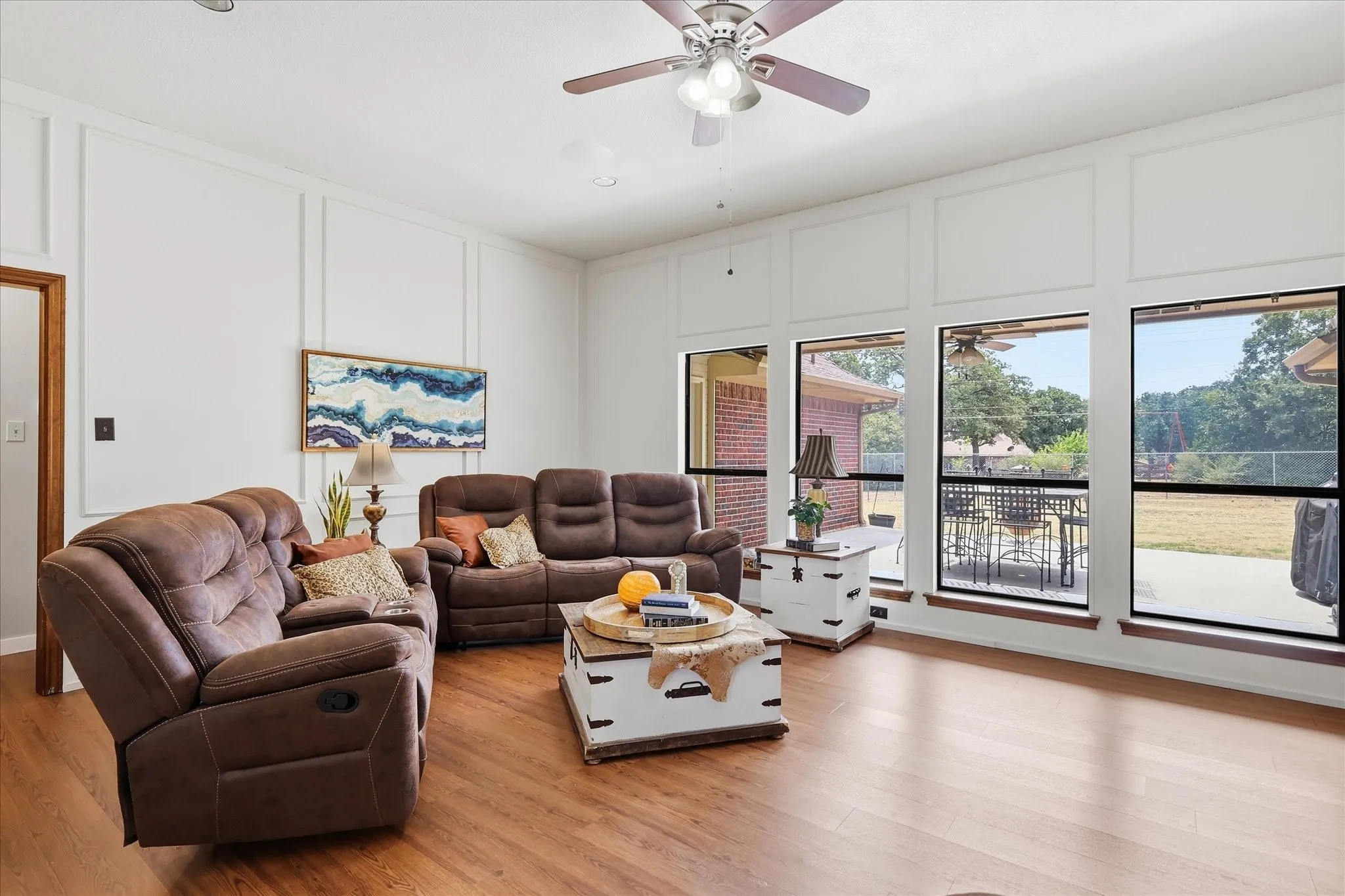 Wood-finish flooring throughout, accent wall paneling, ceiling fan with integrated lighting, and multiple large windows