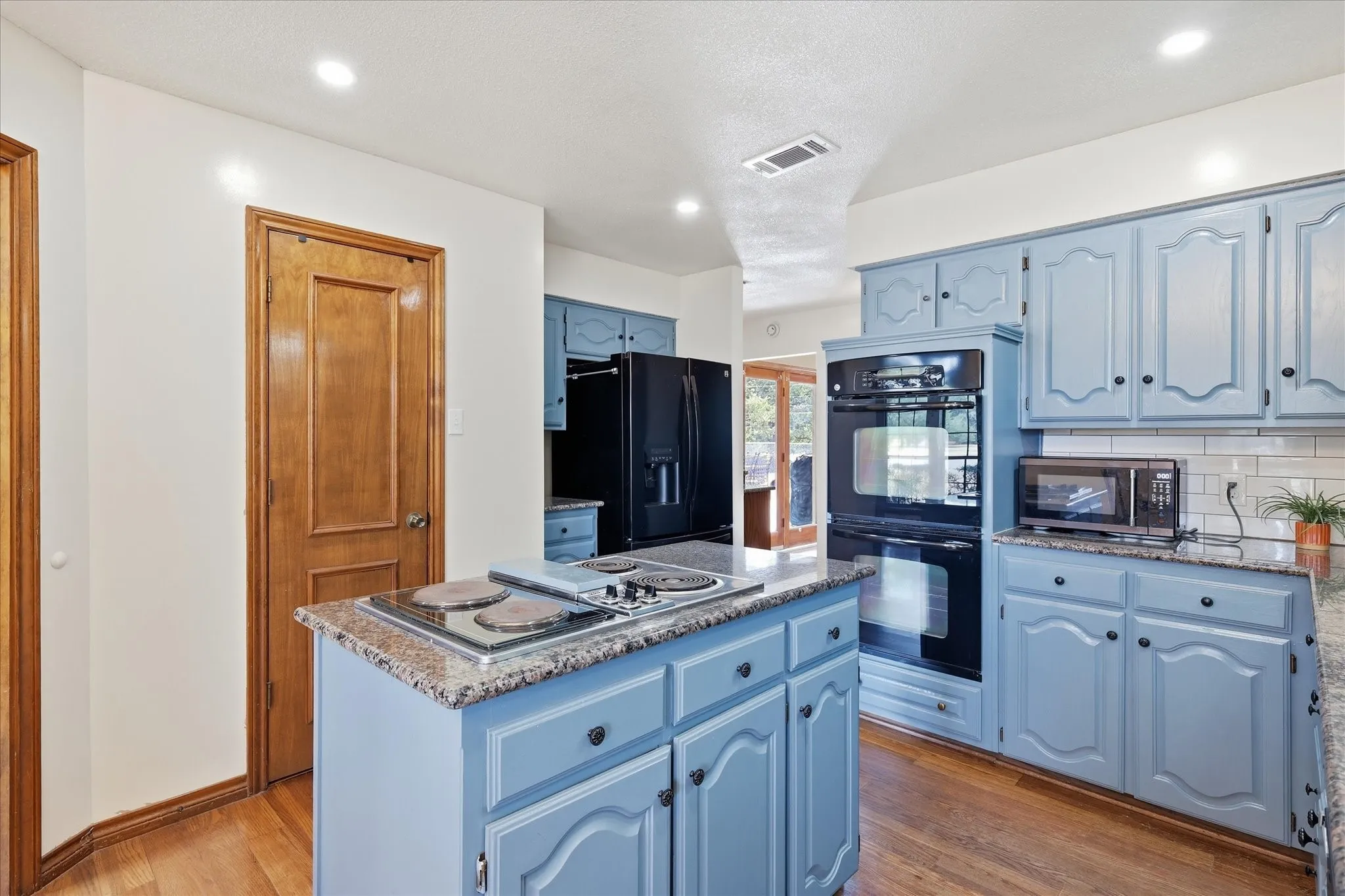 Kitchen featuring wood-finish flooring, light blue cabinetry, granite-finish countertops, built-in double ovens, and a central island with a cooktop