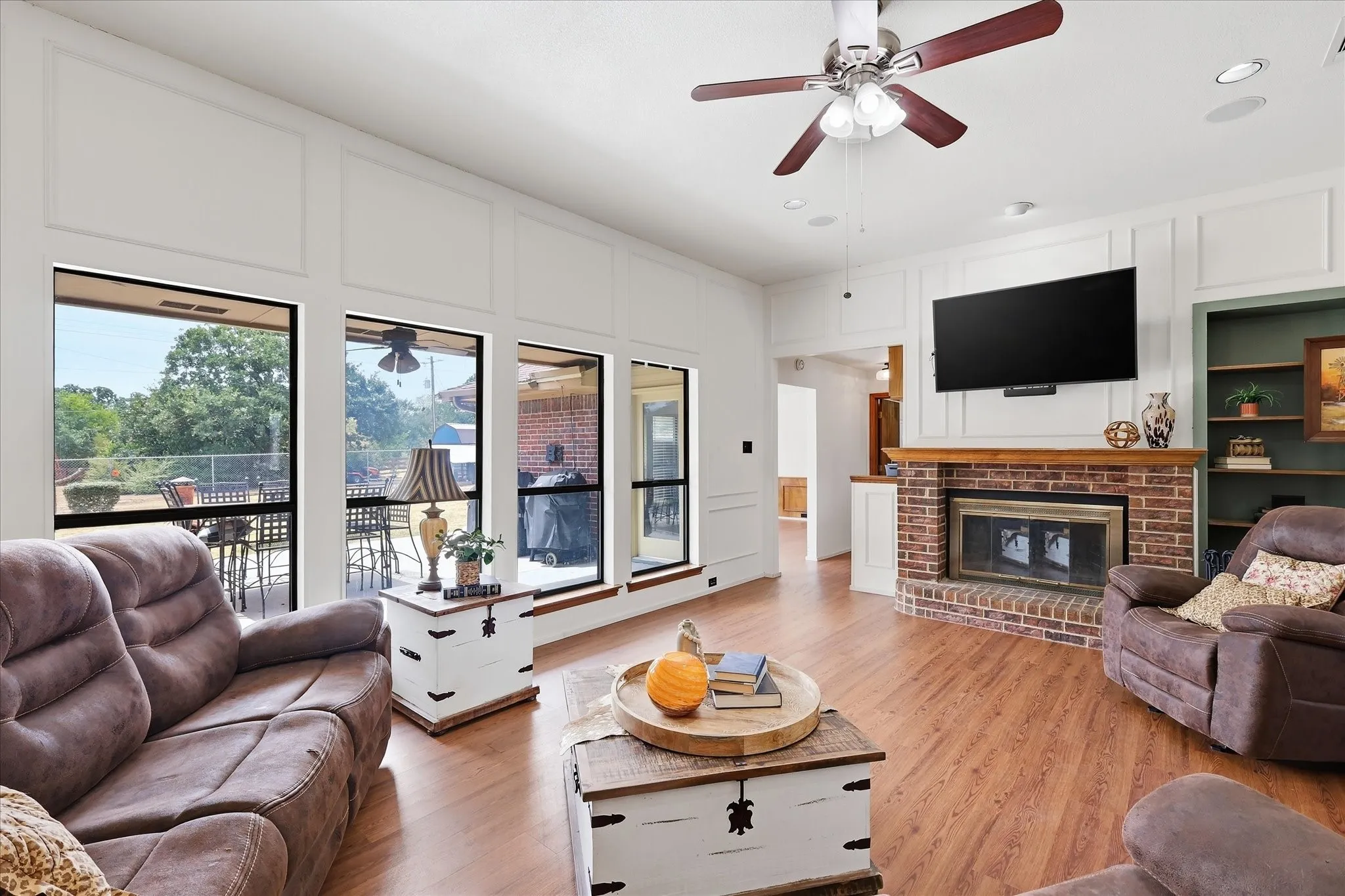 Expansive living area featuring large windows, wood-finish flooring, a brick fireplace with a wood mantel, and a ceiling fan