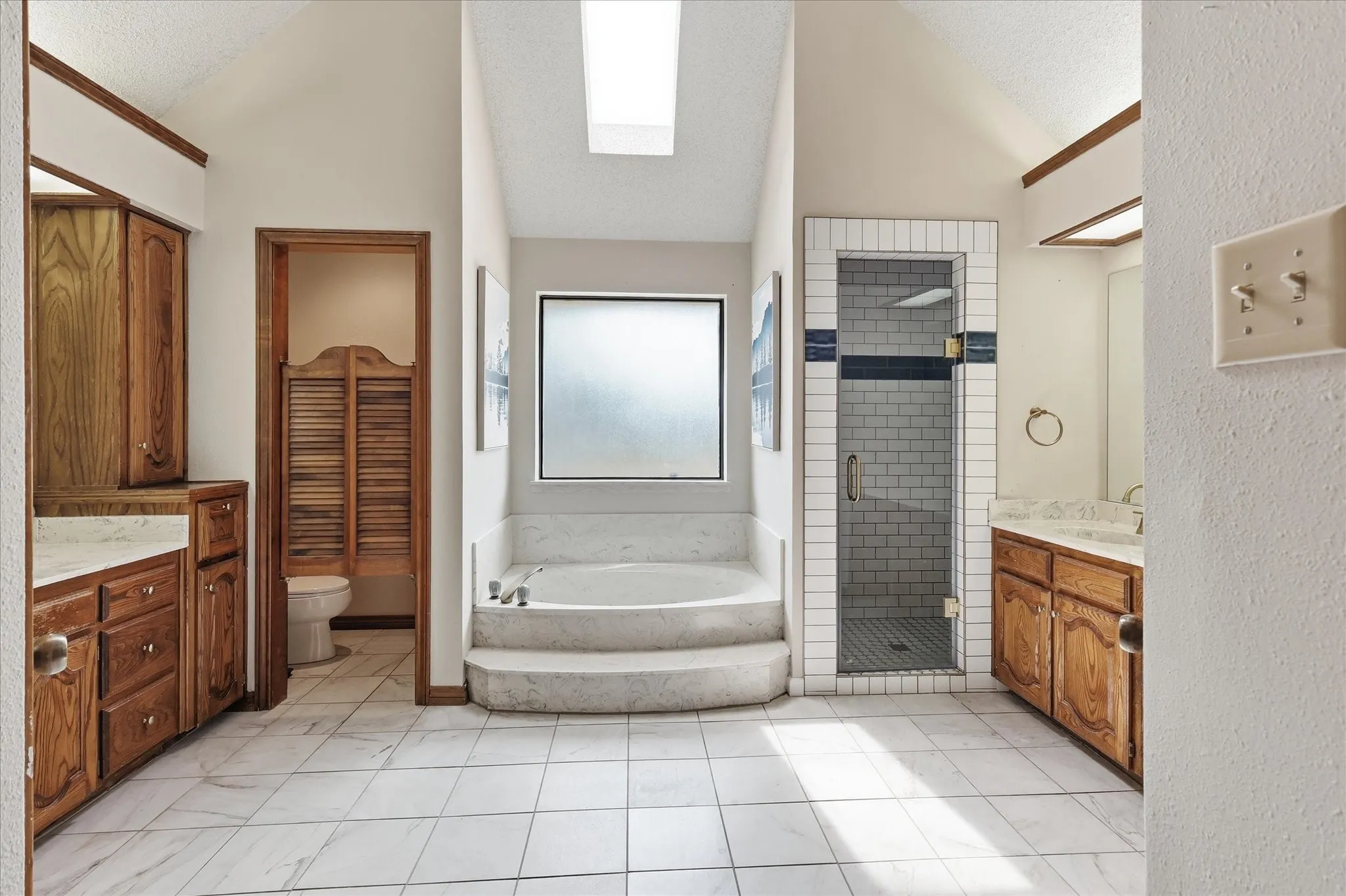 Tiled flooring, built-in soaking tub, glass-enclosed shower, dual wood-finish vanities with countertop, and a skylight