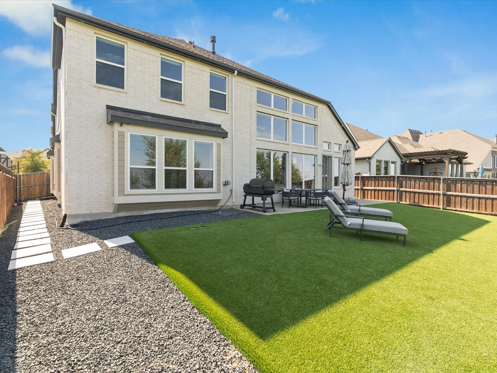 White brick exterior with multiple windows, leading to a fenced backyard featuring artificial turf, a gravel pathway with stepping stones, and a concrete patio