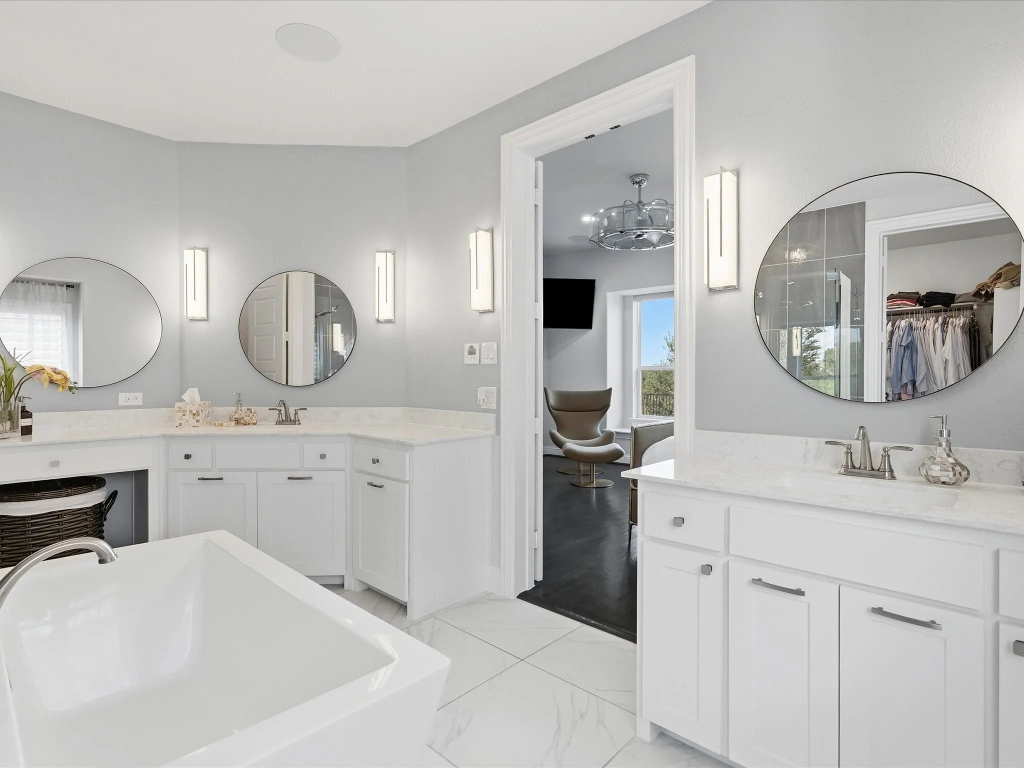 White soaking tub with deck-mounted faucet, dual vanities with round mirrors, and wall-mounted sconces