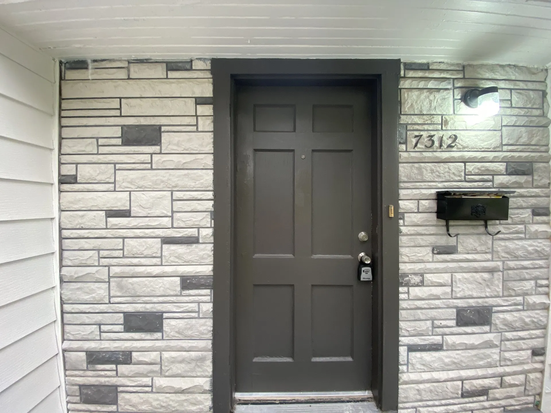 Panel door with dark trim, stone-look siding, and wall-mounted light fixture