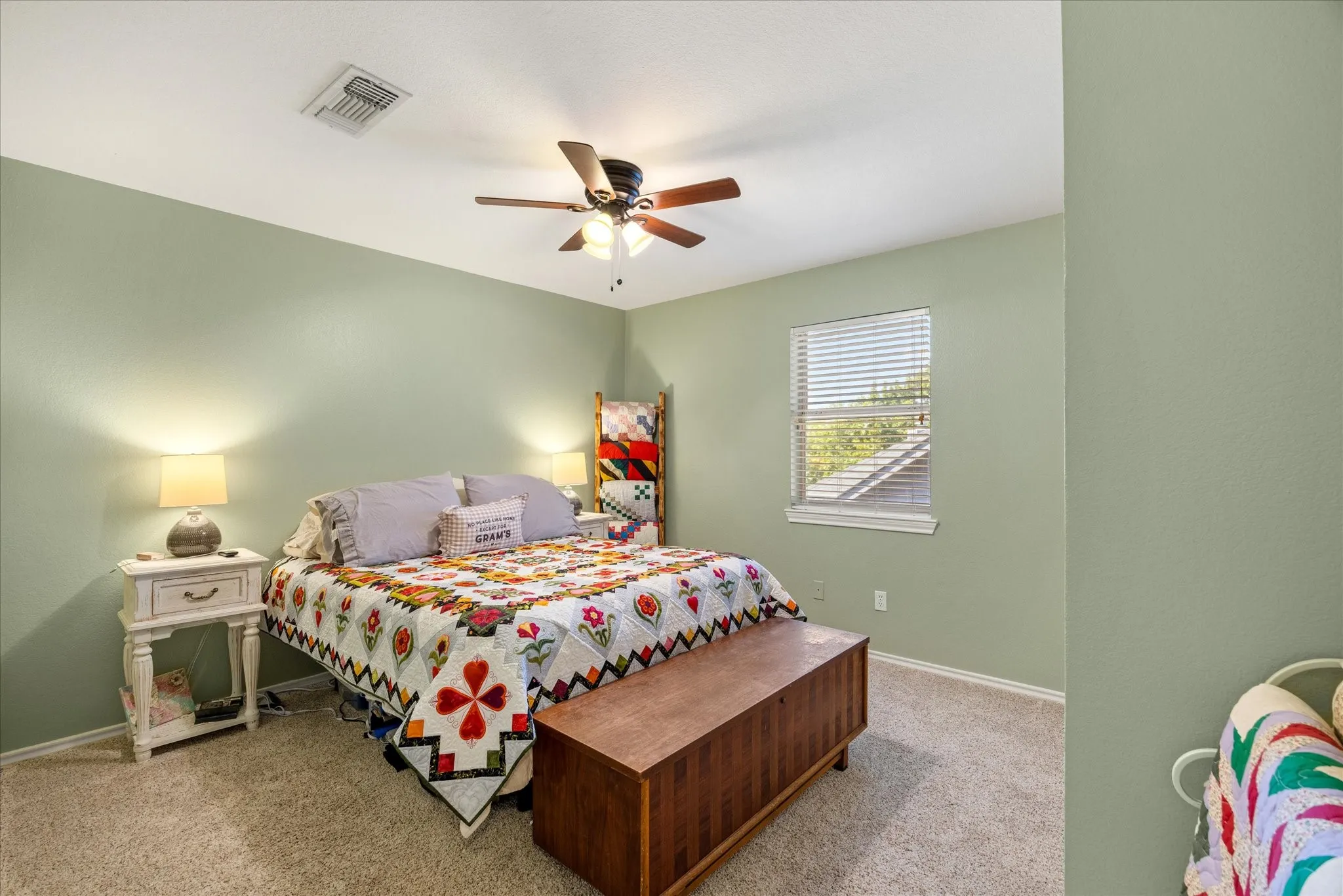 Serene secondary bedroom featuring soft sage-green walls, carpeted flooring, ceiling fan with integrated lighting, two windows with plantation shutters providing natural light, and a wood-finish storage bench at the foot of the bed