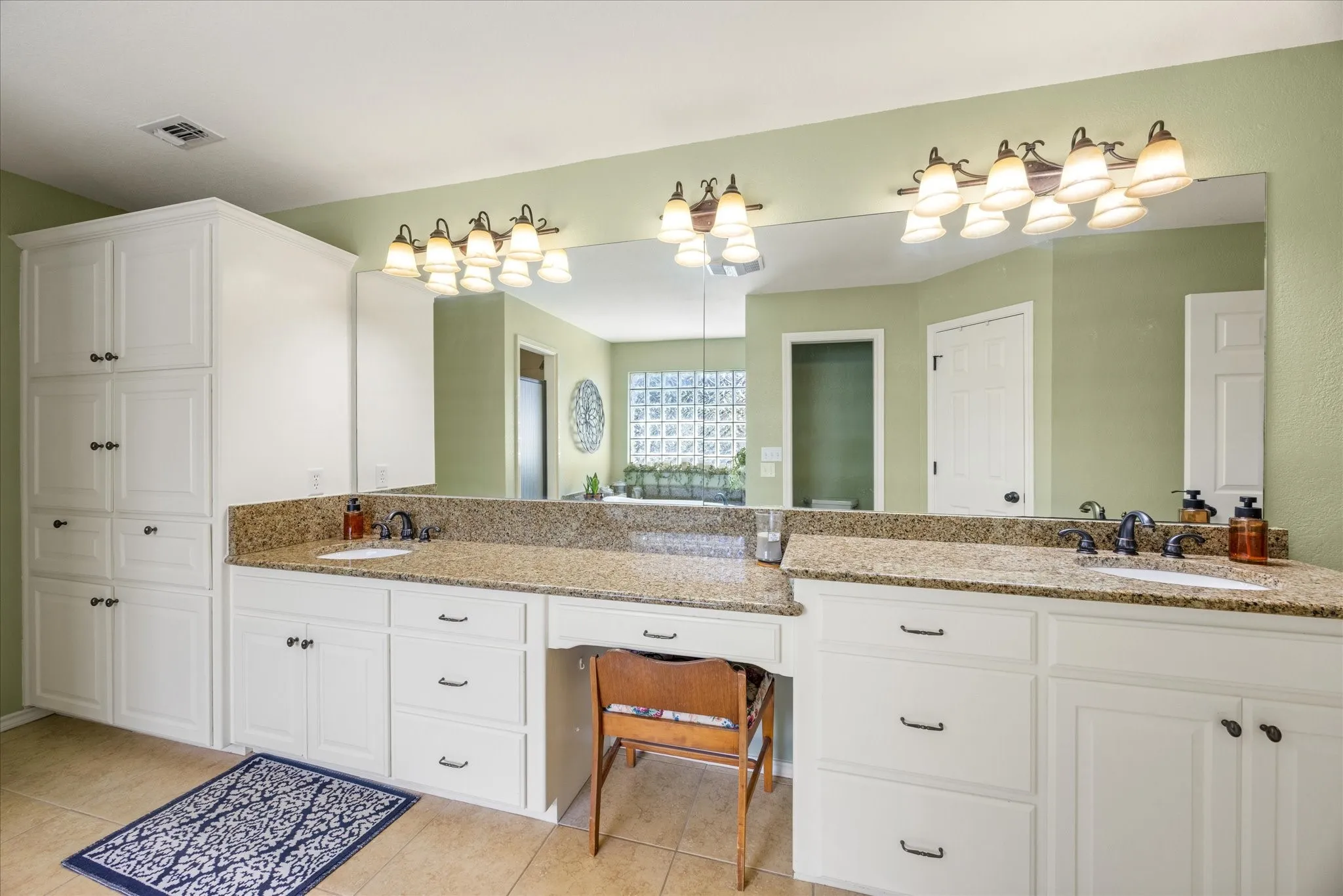 Spa-inspired primary bathroom featuring dual vanities with granite countertops, abundant natural lighting from large mirror surround, chrome hardware and fixtures, white cabinetry with ample storage, and tile flooring throughout
