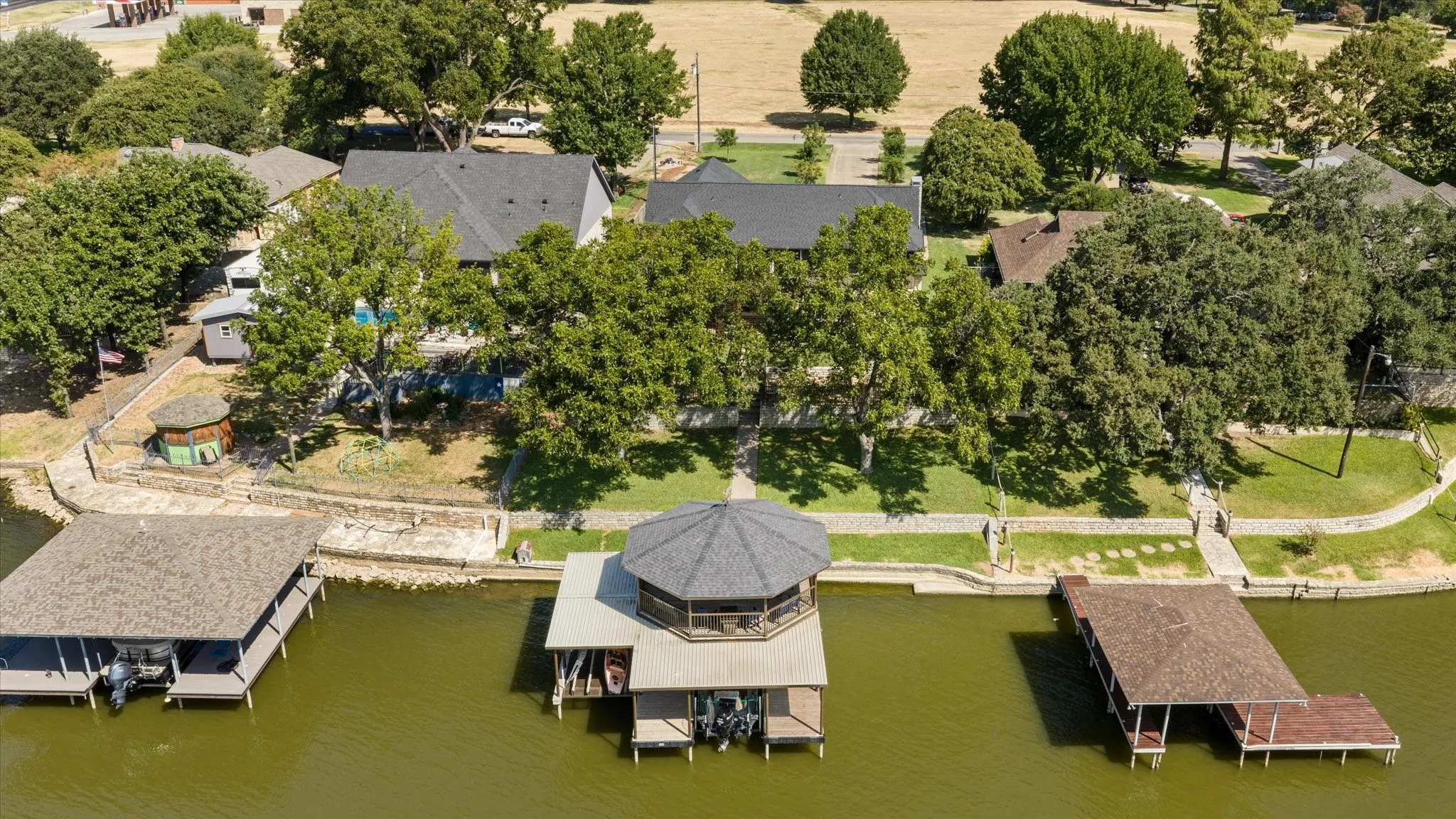 Waterfront property featuring multiple boat docks with covered structures, expansive manicured grounds anchored by mature shade trees, and a main residence with dark-shaded roofing visible in the background