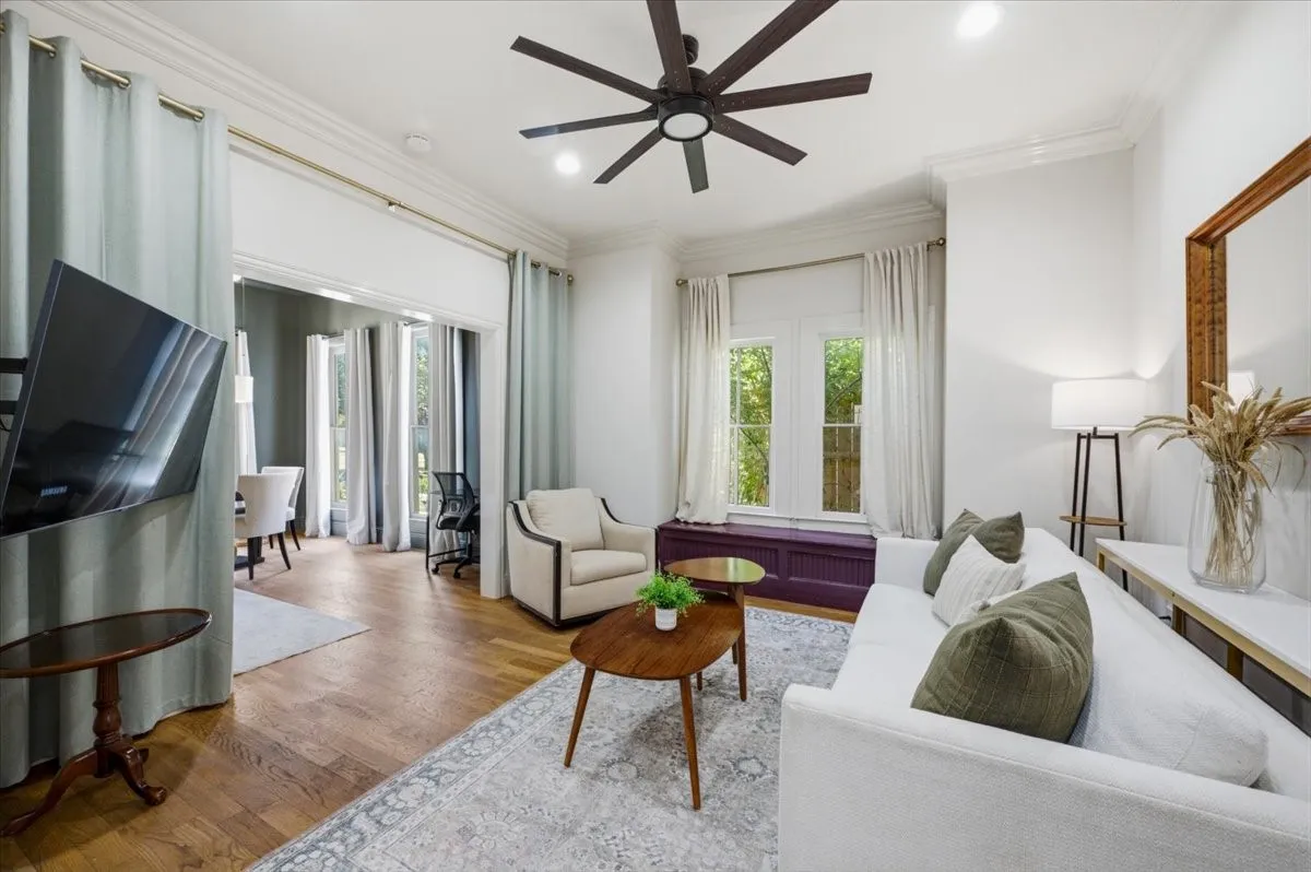 Wood-finish flooring, crown molding, a dark-toned ceiling fan, and large windows with privacy coverings