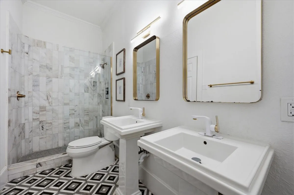 Dual pedestal sinks with gold-framed mirrors, a walk-in shower with vertical marble-look tile, and patterned floor tile