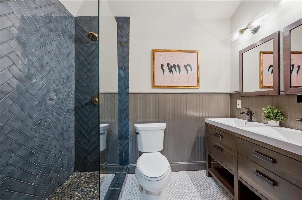 Herringbone tile shower with pebble floor, glass shower enclosure, dual vanity with undermount sinks, and beadboard wainscoting