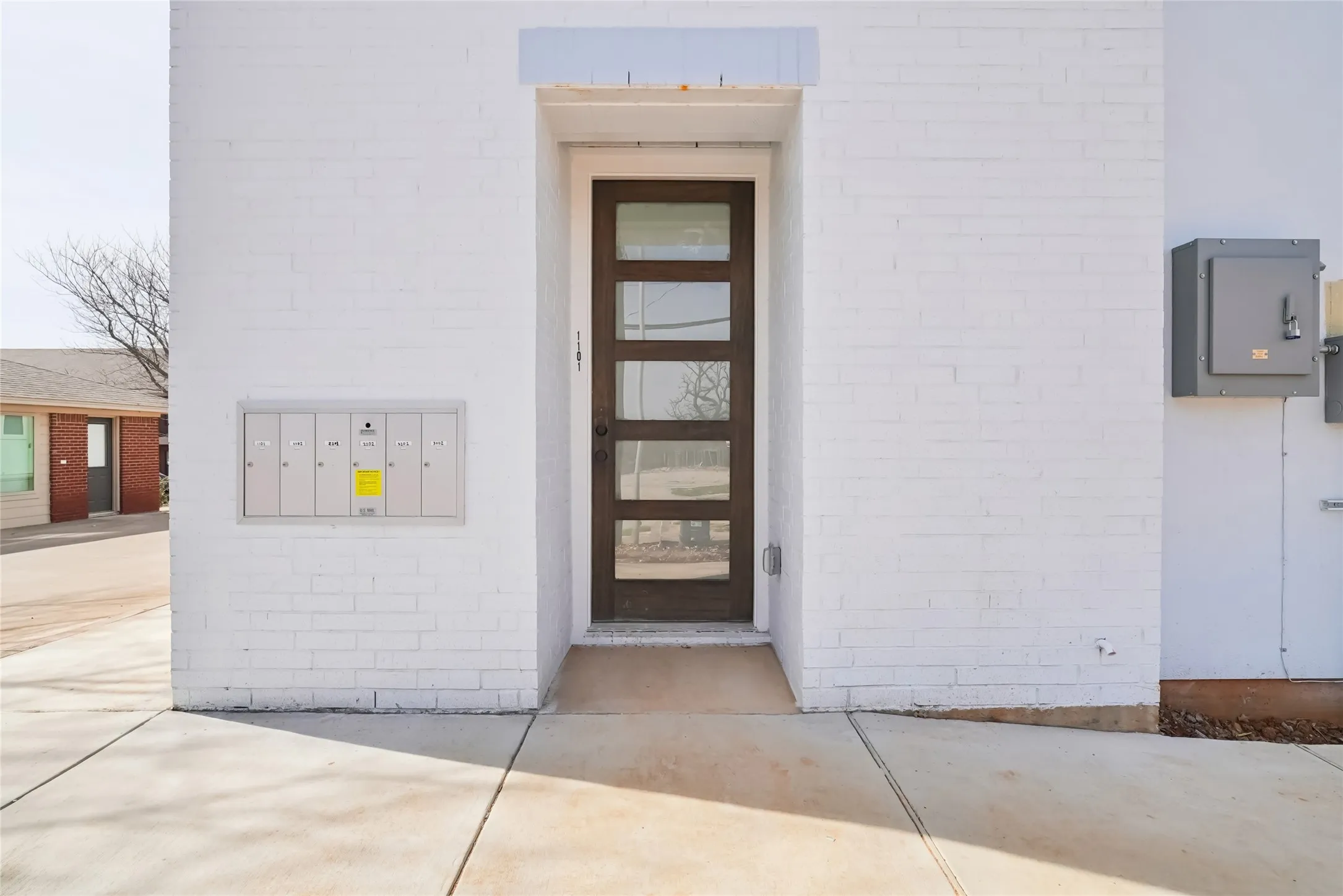 White brick exterior with a modern entry door featuring multiple glass panels, an integrated mail collection unit, and a concrete pathway