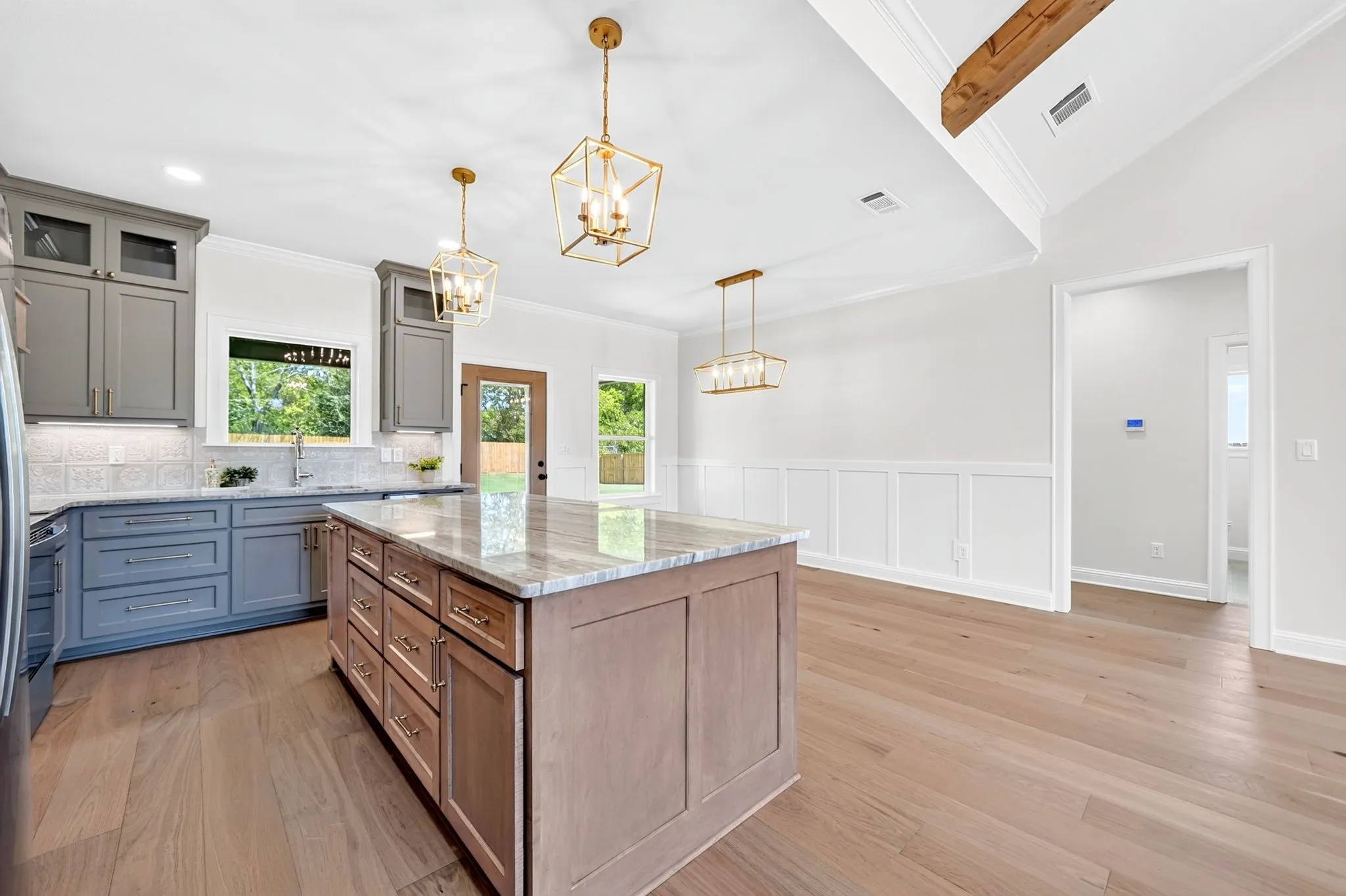 Two-tone kitchen cabinetry, wood-finish flooring, gold-tone pendant lighting, exposed wood beam, and wainscoting details