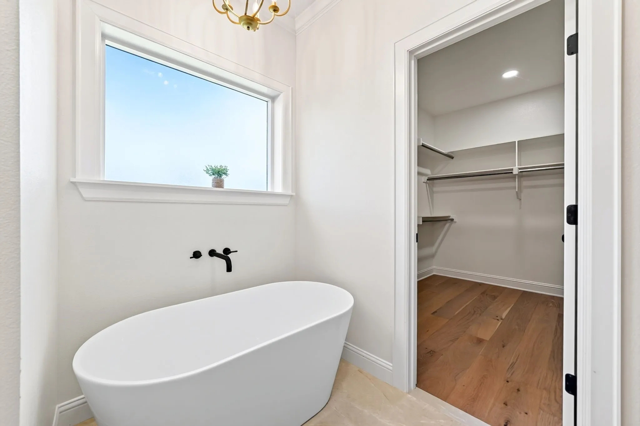 Freestanding oval soaking tub with matte black wall-mounted faucet, large window, and light tile flooring
