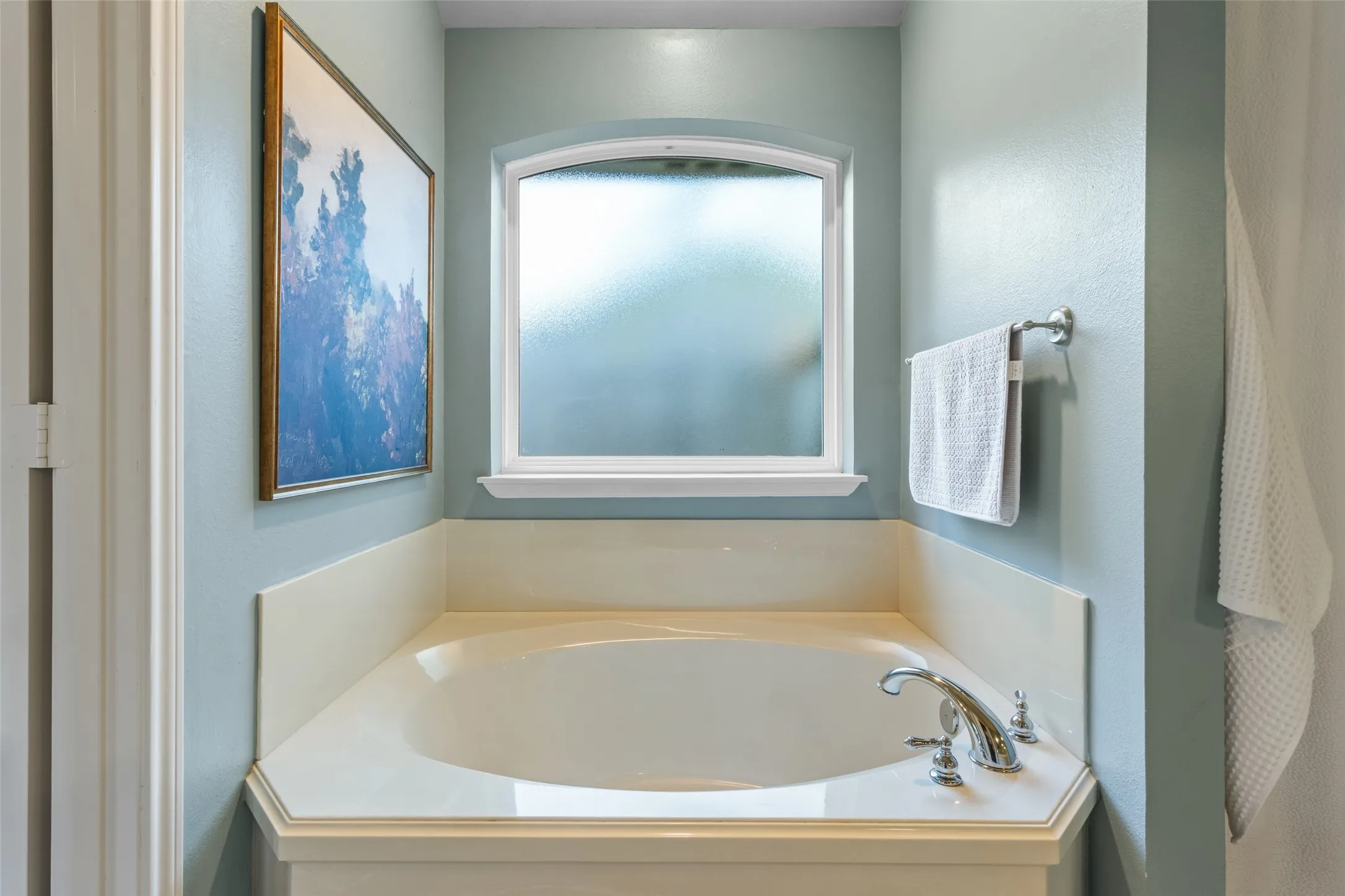 Built-in corner soaking tub with deck-mounted chrome fixtures, an arched frosted window, and a surrounding light-toned ledge