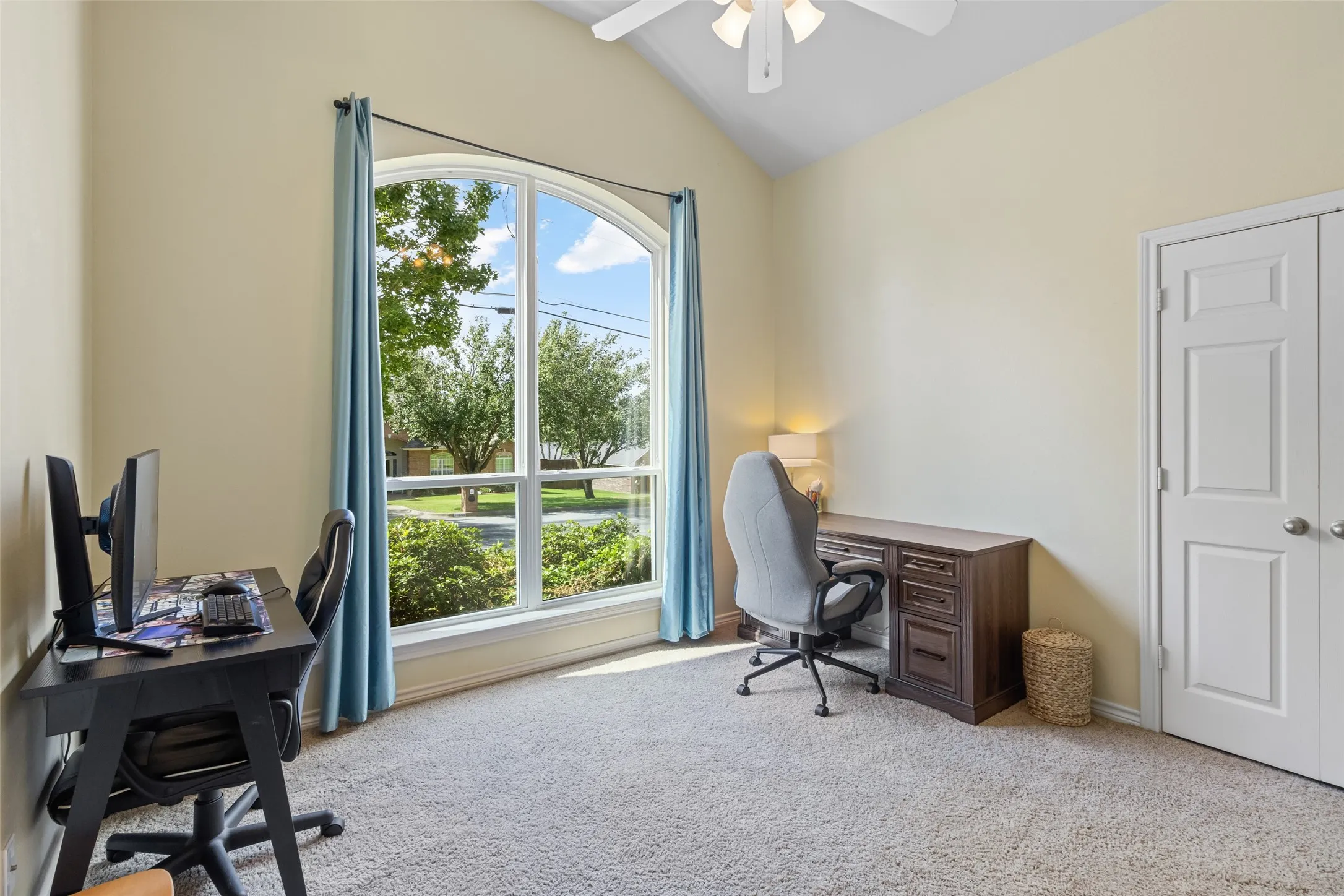 Bright, naturally lit home office featuring expansive glass French doors framed in blue, gleaming wood-finish desk with integrated storage, ergonomic office seating, neutral carpet flooring, and a modern ceiling fan