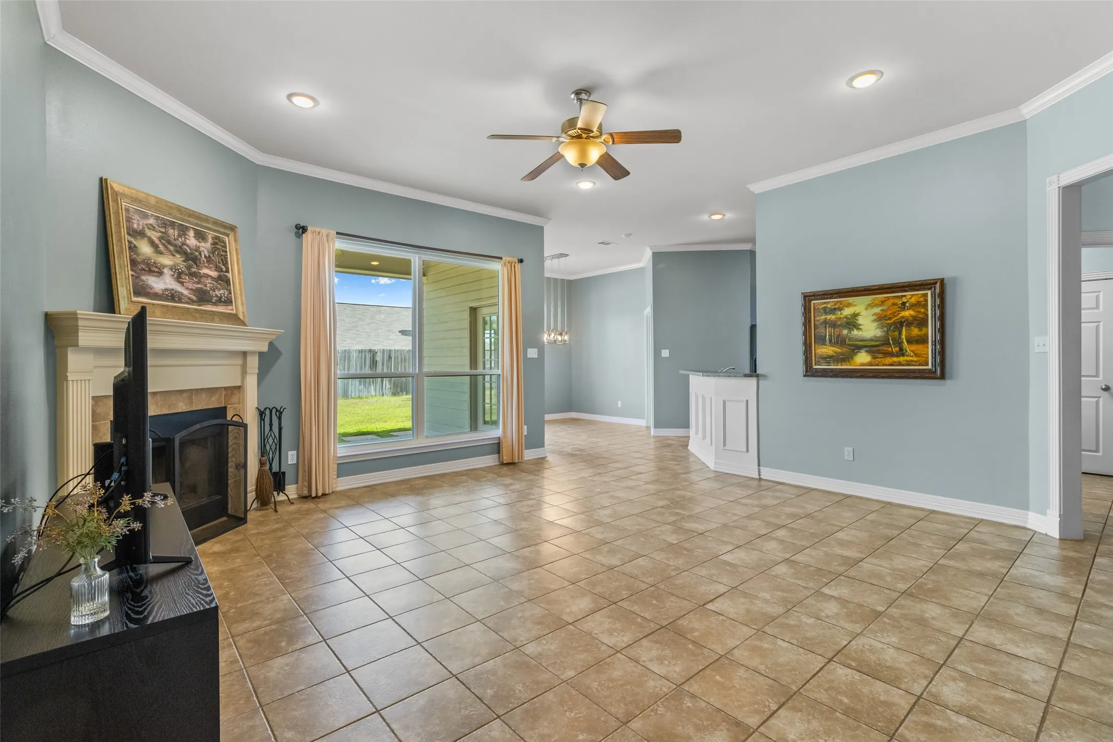 Spacious living area featuring tile flooring, a fireplace with a mantel, a ceiling fan, recessed lighting, and a large window overlooking the backyard