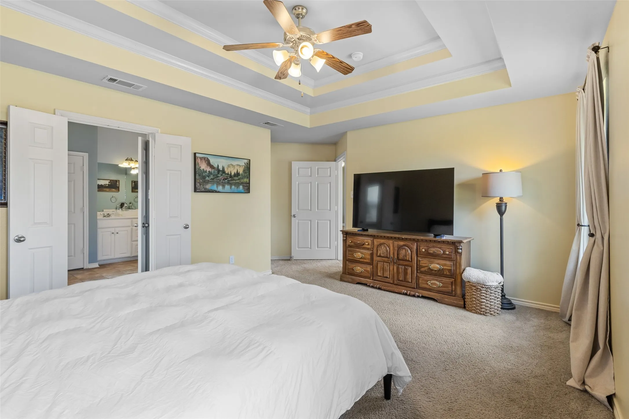 Spacious master suite featuring a tray ceiling with ceiling fan, soft yellow walls, and neutral carpeting