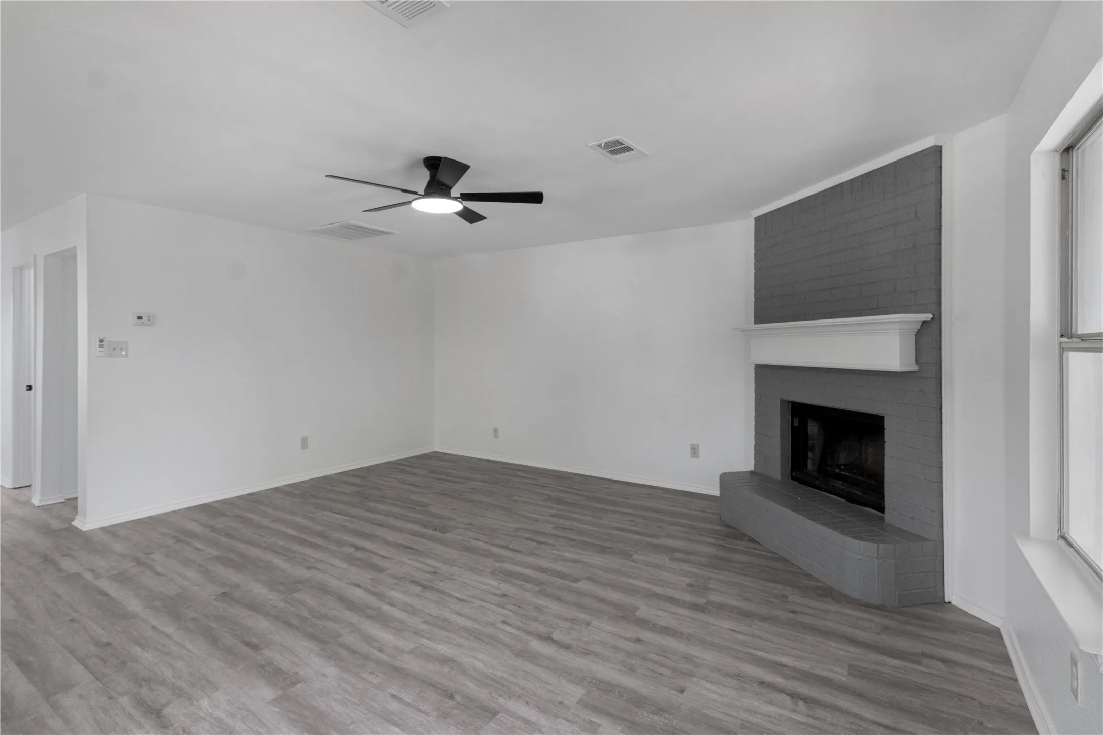 Spacious room featuring wood-finish flooring, a contemporary ceiling fan, and a brick fireplace with a white mantel and hearth