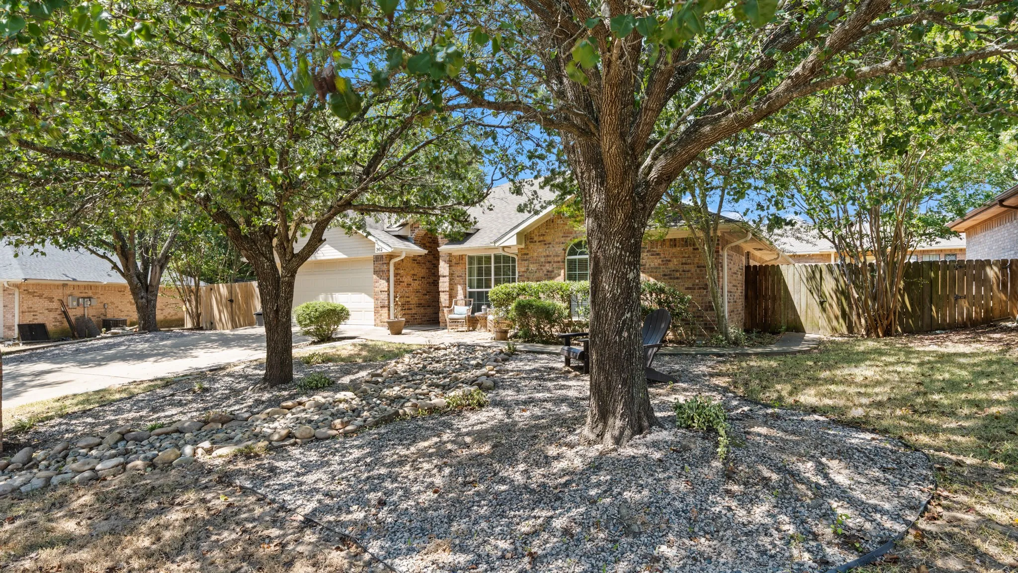 Brick exterior, attached garage, mature trees, gravel landscaping, and a fenced yard