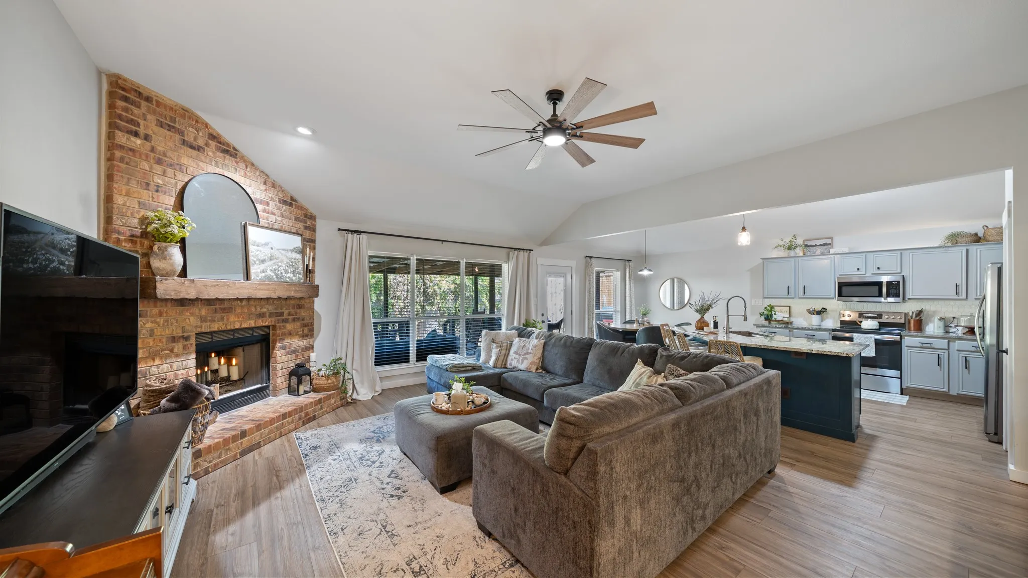 Vaulted ceiling with a ceiling fan, brick fireplace, wood-finish flooring, and light-toned cabinetry with an island