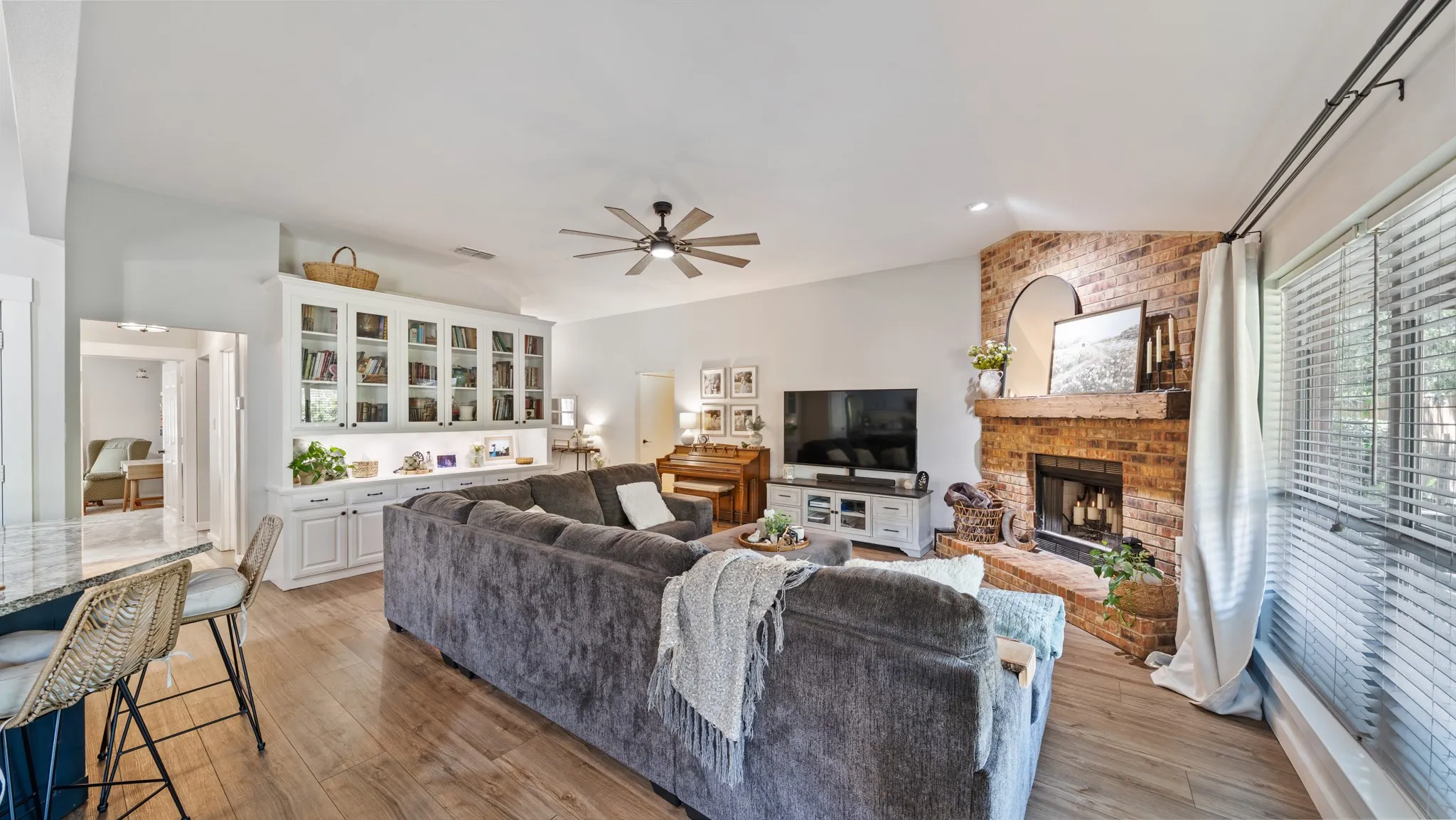 Wood-finish flooring, built-in white cabinetry with glass fronts, a corner brick fireplace, a ceiling fan, and large windows with blinds