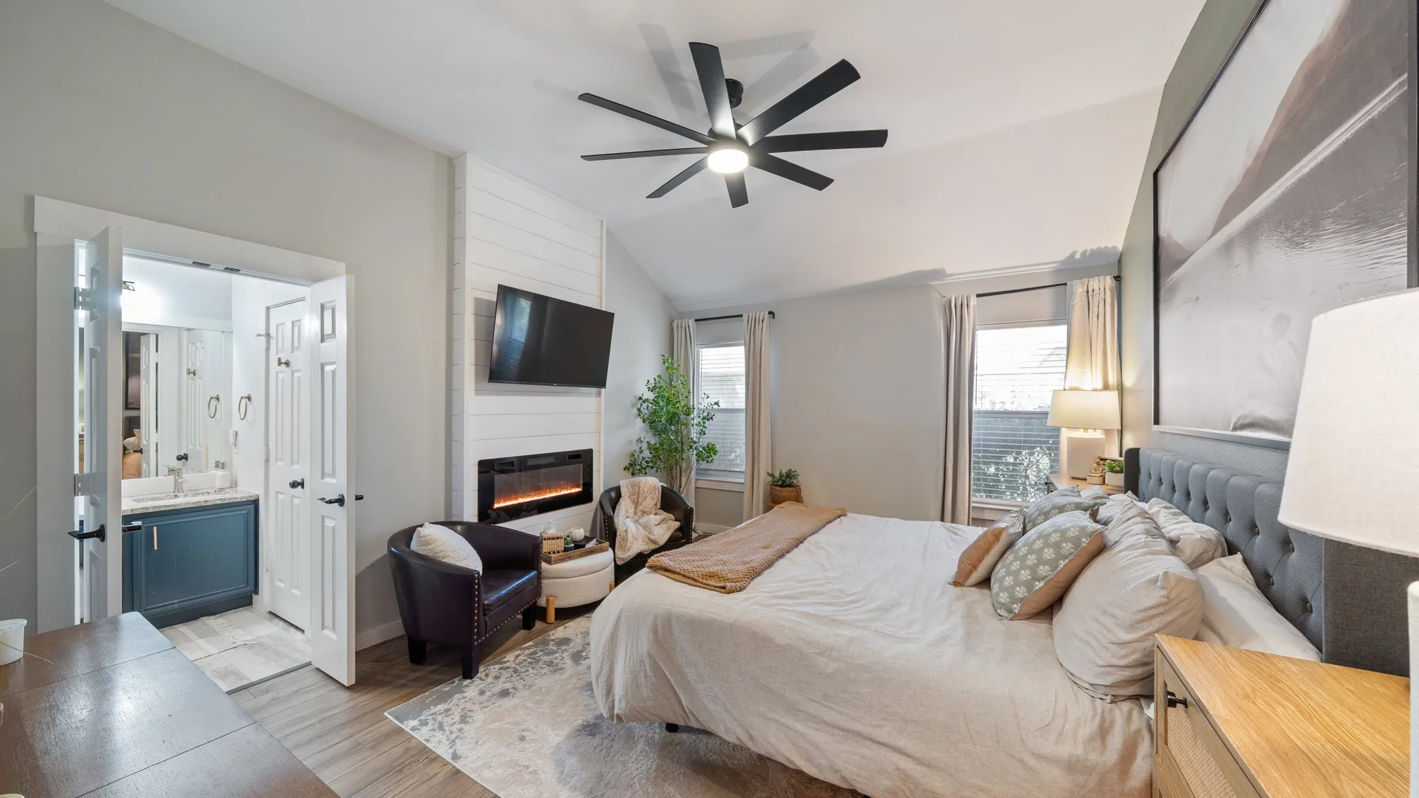 Vaulted ceiling with a contemporary ceiling fan, shiplap accent wall with an electric fireplace, wood-finish flooring, and two windows with blinds