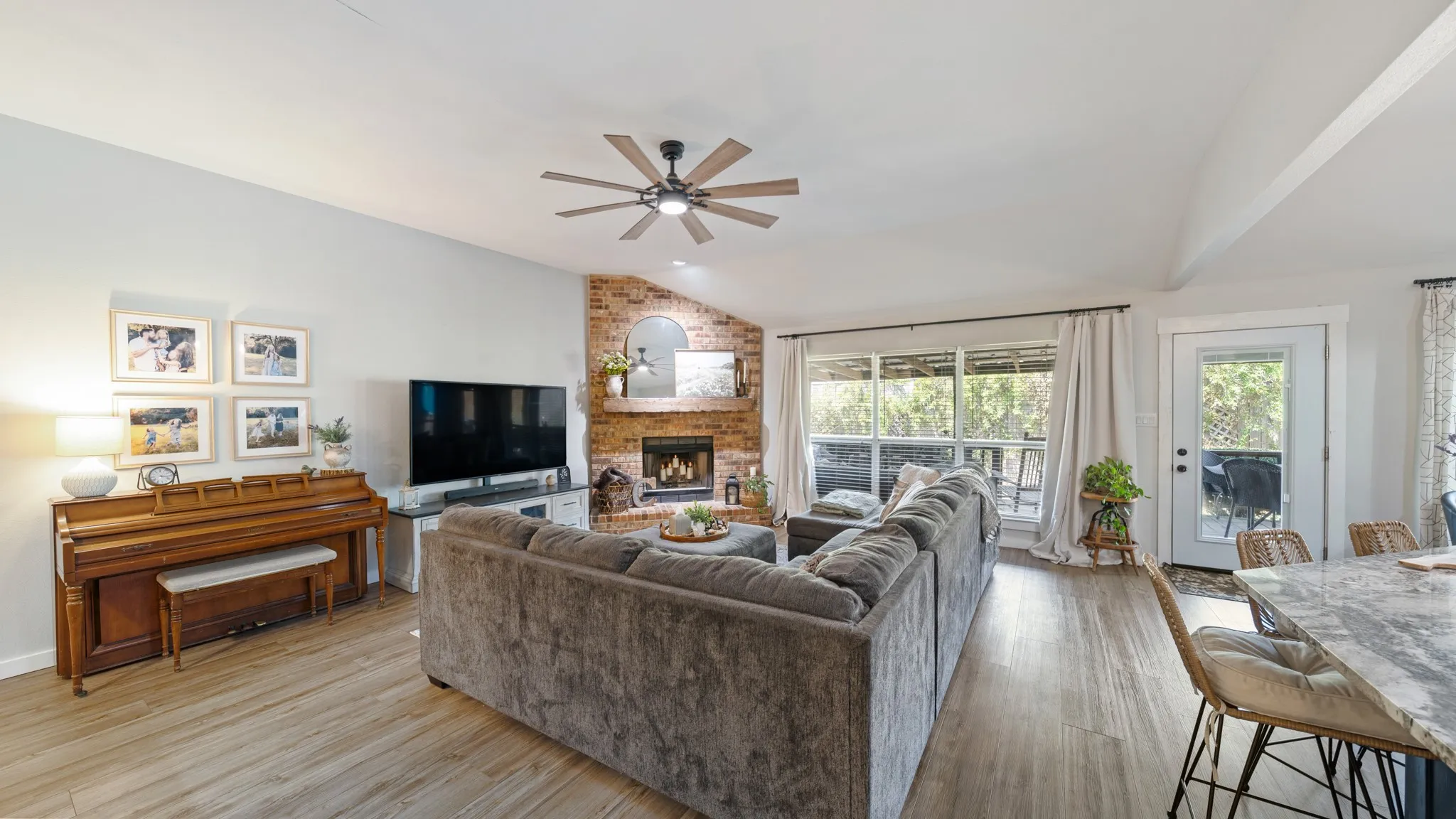 Vaulted ceiling with a ceiling fan, brick fireplace, wood-finish flooring, large windows, and a glass-paneled door
