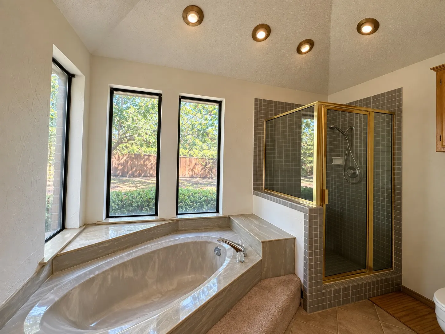 Soaking tub with a tiled surround, three large windows, a separate shower with a glass enclosure, and recessed ceiling lighting