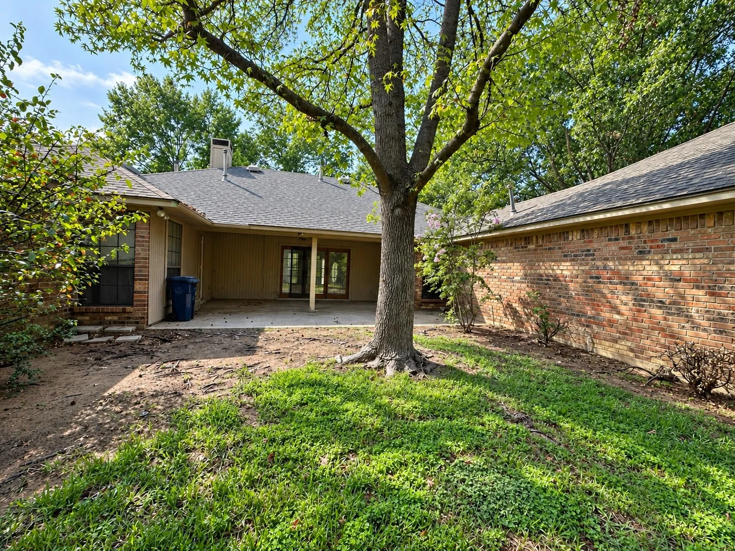 Patio framed by mature trees, a wooden fence, and lush landscaping, leading to a garage with a panel door